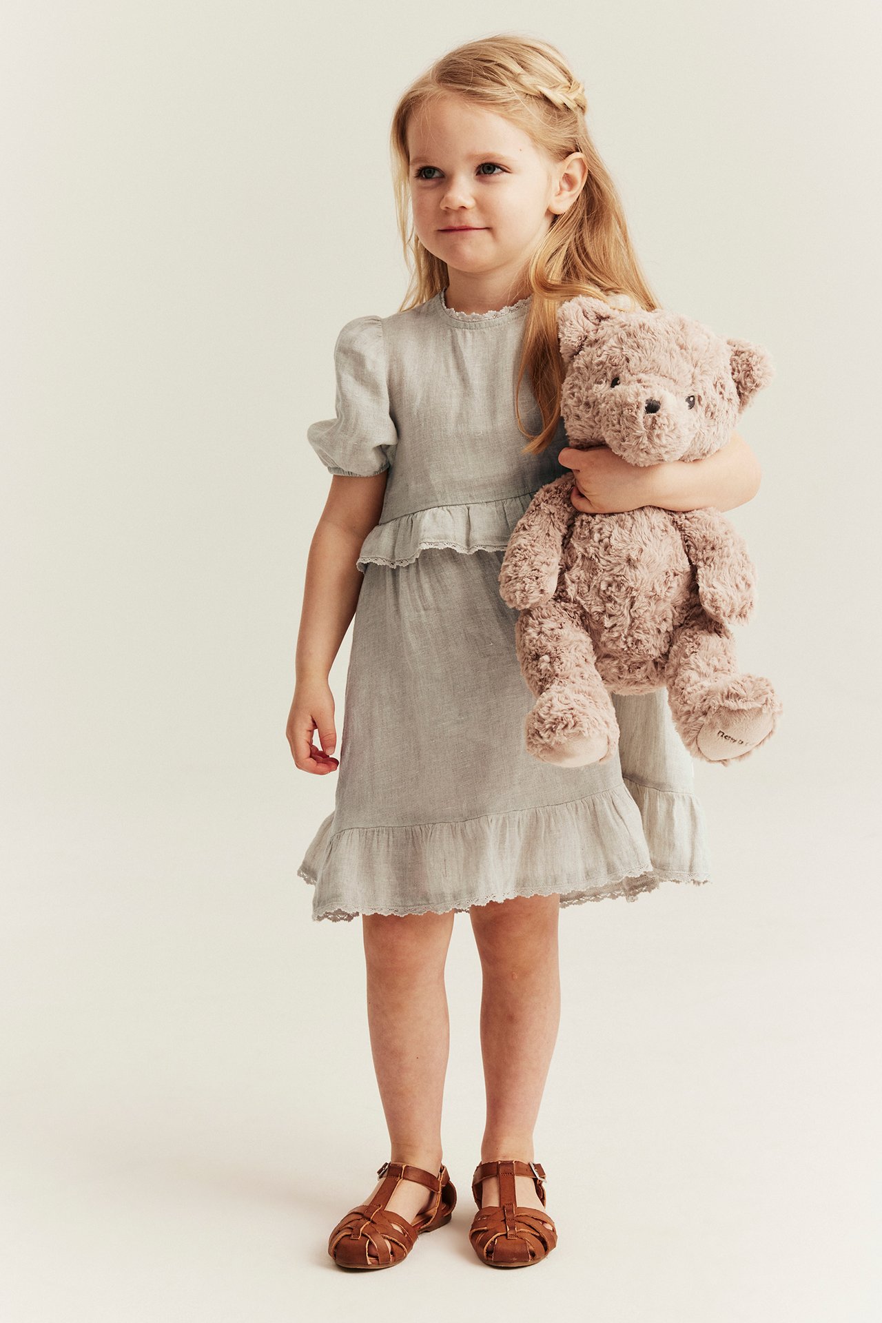 Kid in a light beige linen dress with puff sleeves and ruffles, holding a brown teddy bear and brown sandals.