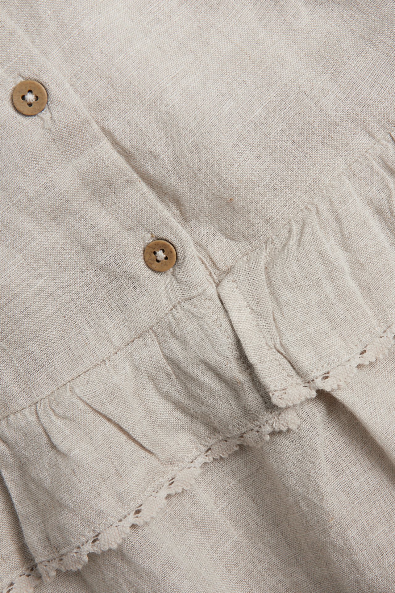 Nahaufnahme eines hellbeigen Leinenkleides mit Rüschen, Spitzenborte und zwei braunen Holzknöpfen.