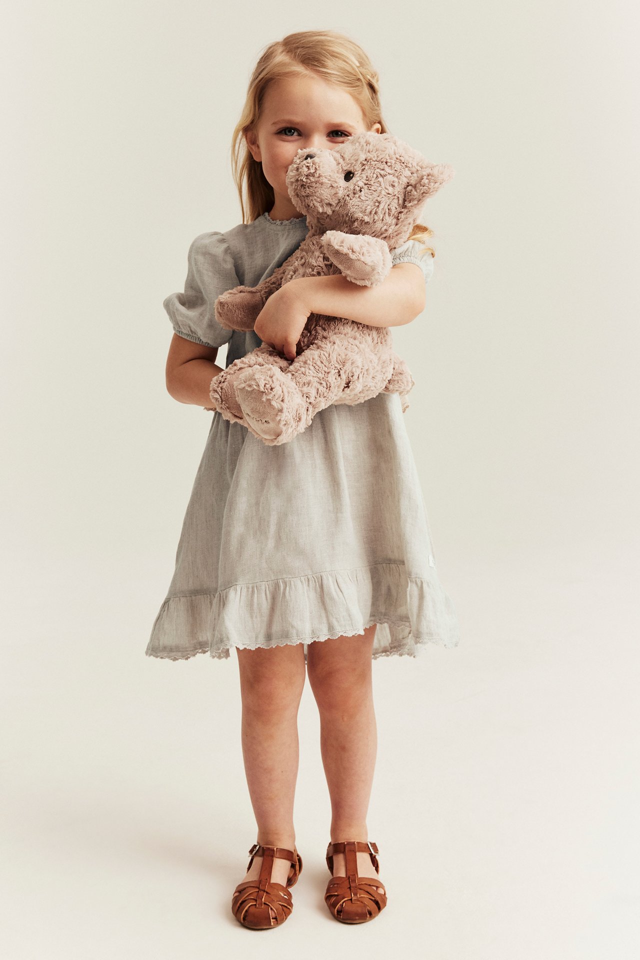 Front view: Kid in light beige linen ruffle dress with puff sleeves, brown sandals, holding teddy bear.