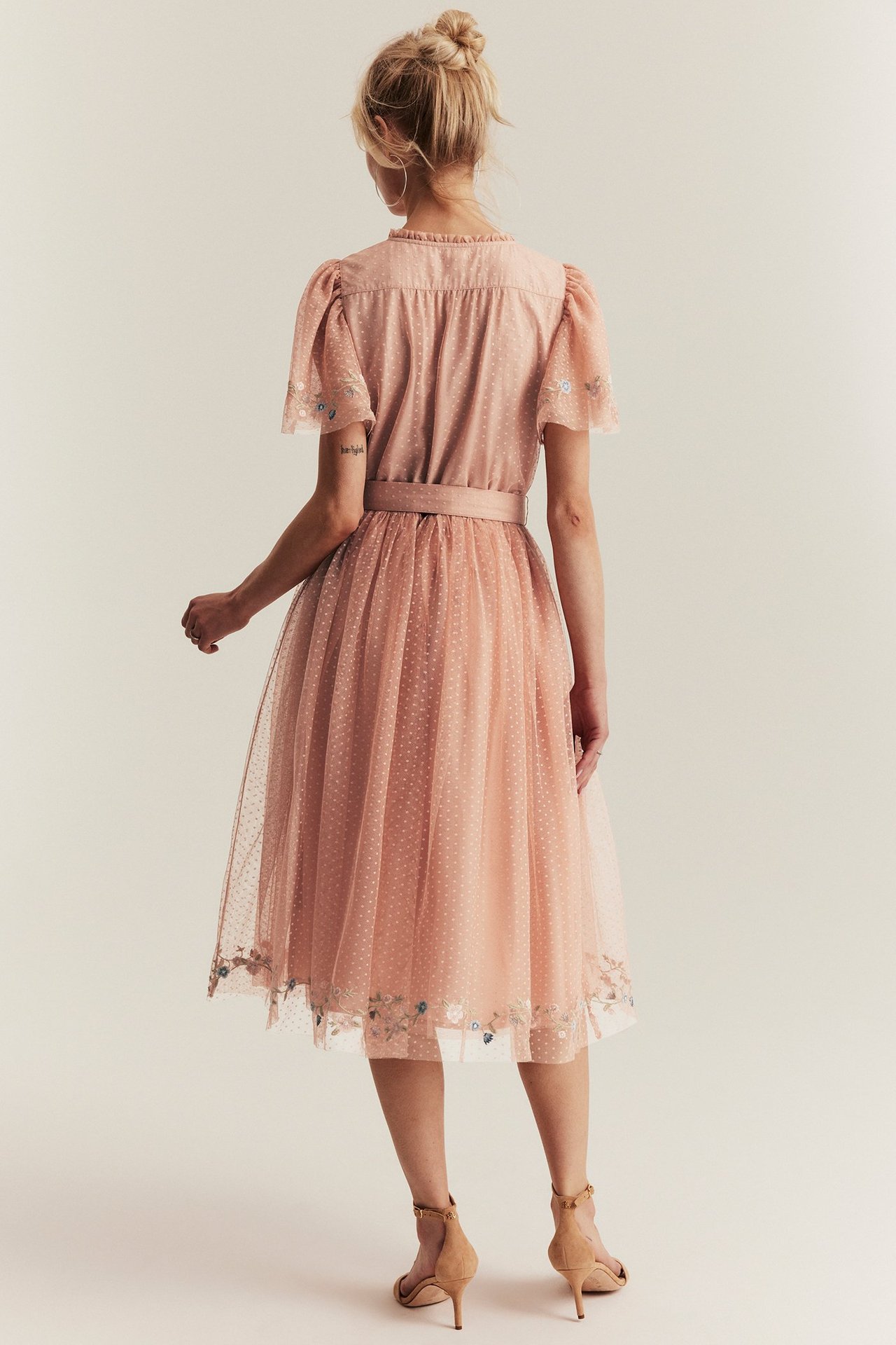 Rückenansicht einer Frau in einem rosa gepunkteten Netz-Midikleid mit Blumenstickerei und beigen Sandaletten.