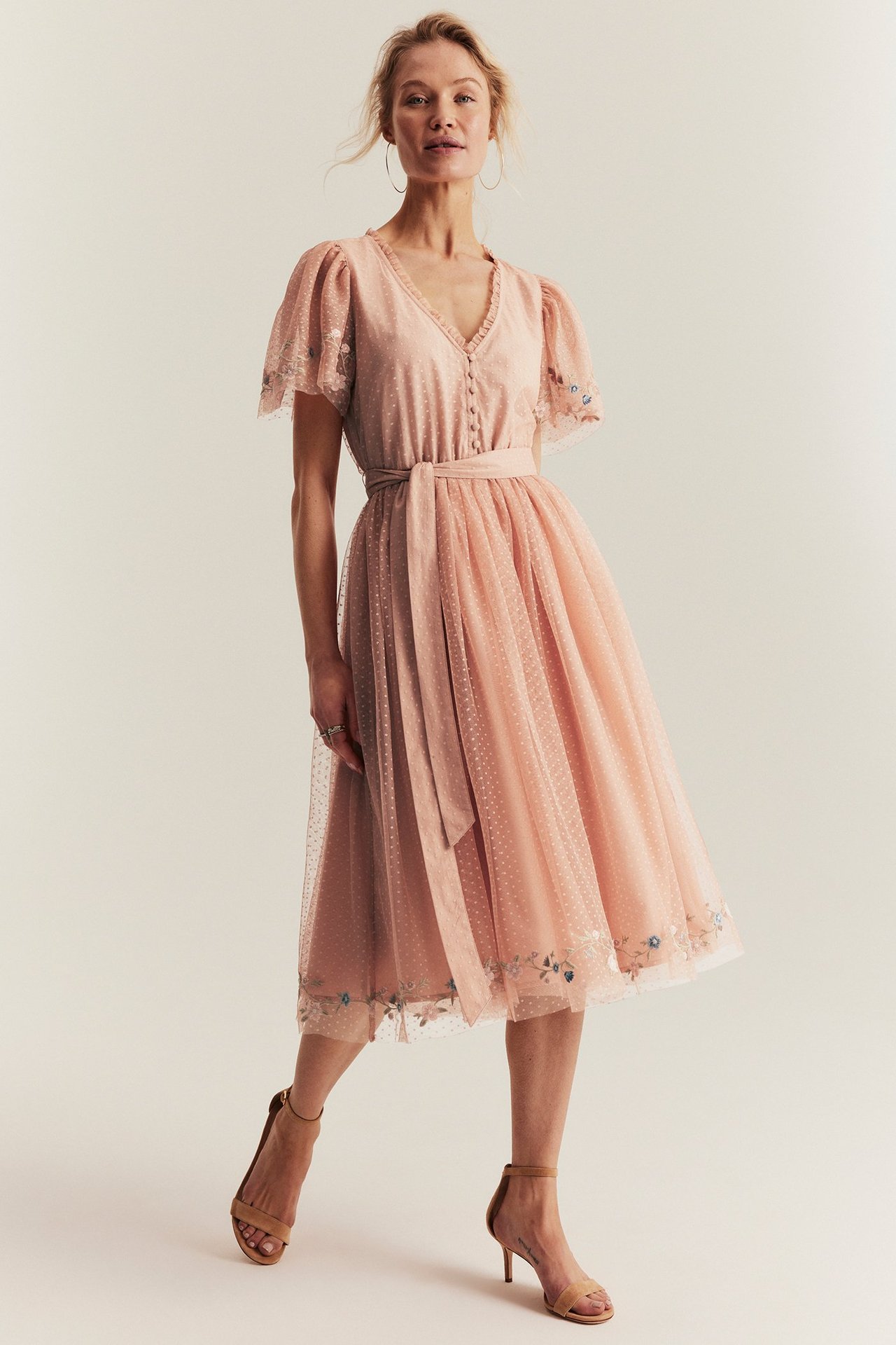 Front view of a woman wearing a pink dotted mesh dress with floral embroidery, V-neck, and tan sandals.