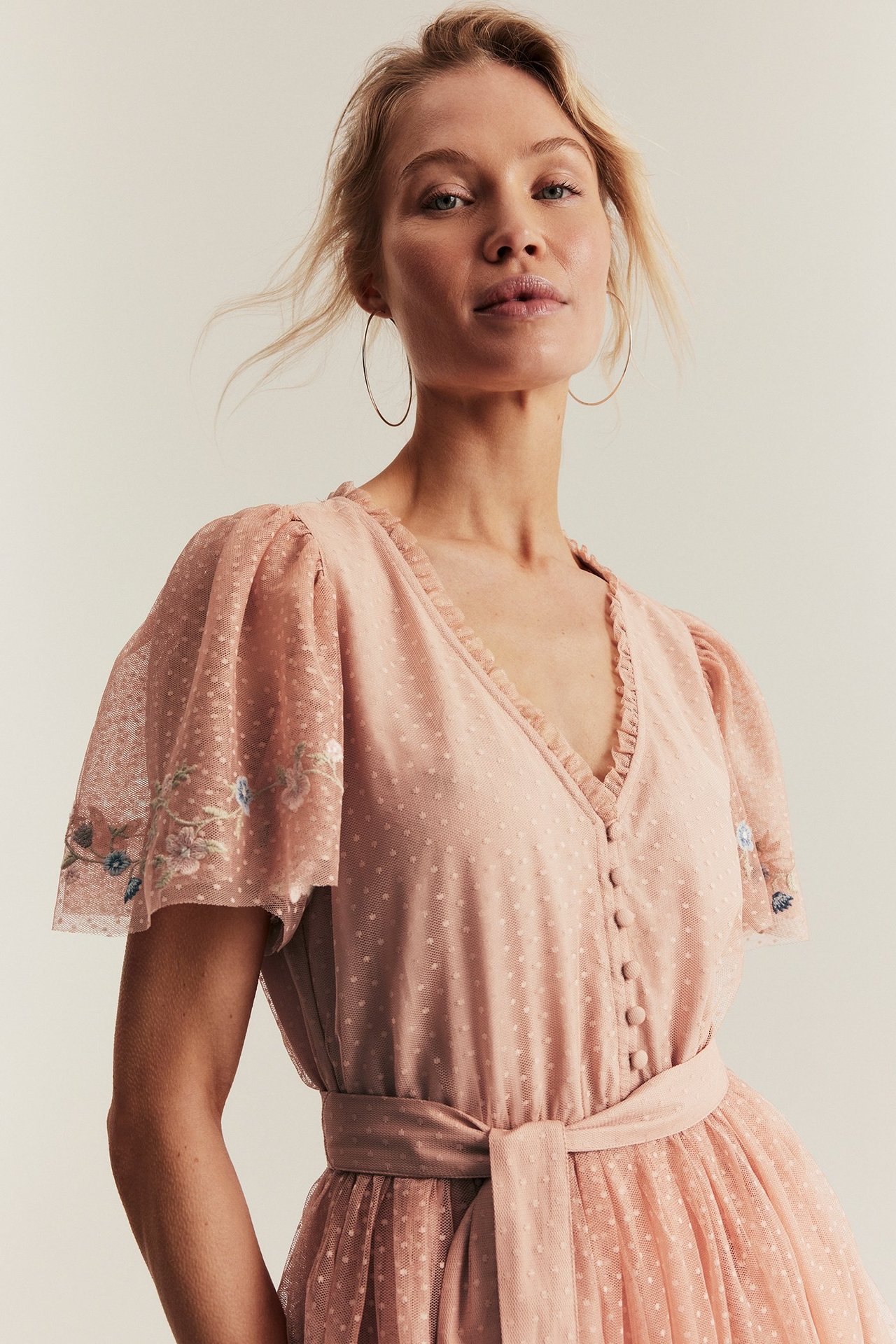 Front view: Woman in pink dotted mesh dress with embroidered sleeves, front buttons, and tied belt.