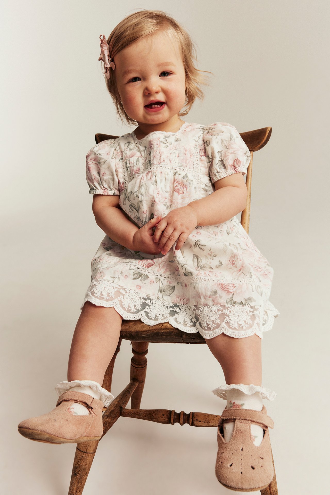 Front view: baby in offwhite floral puff-sleeve dress with lace, pink hair clip, white socks, brown shoes.