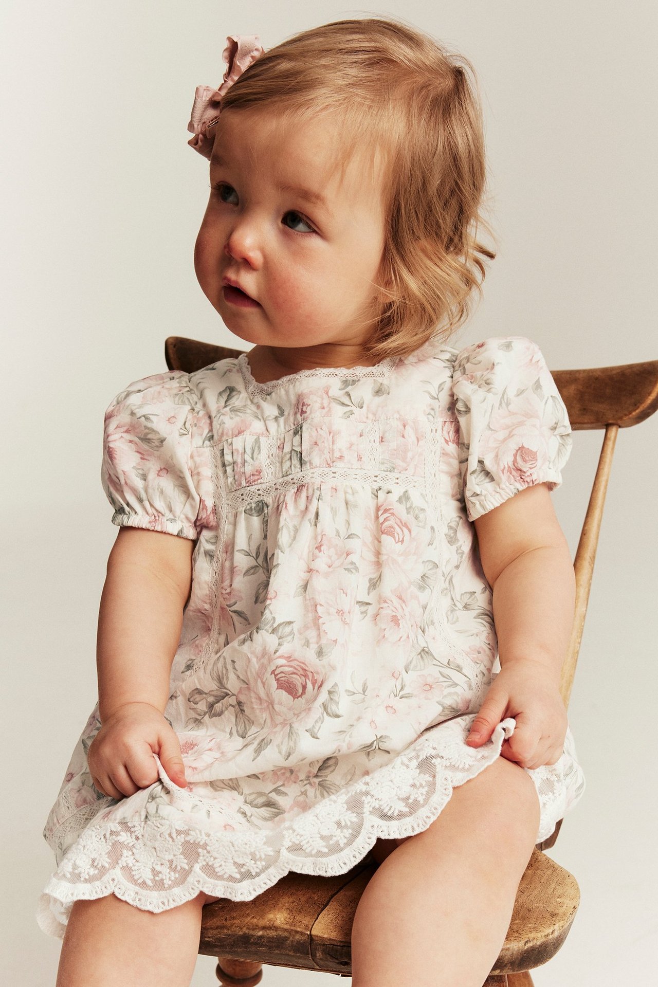 Side view of baby in off-white floral cotton dress with puff sleeves, lace trim, and a pink hair bow.