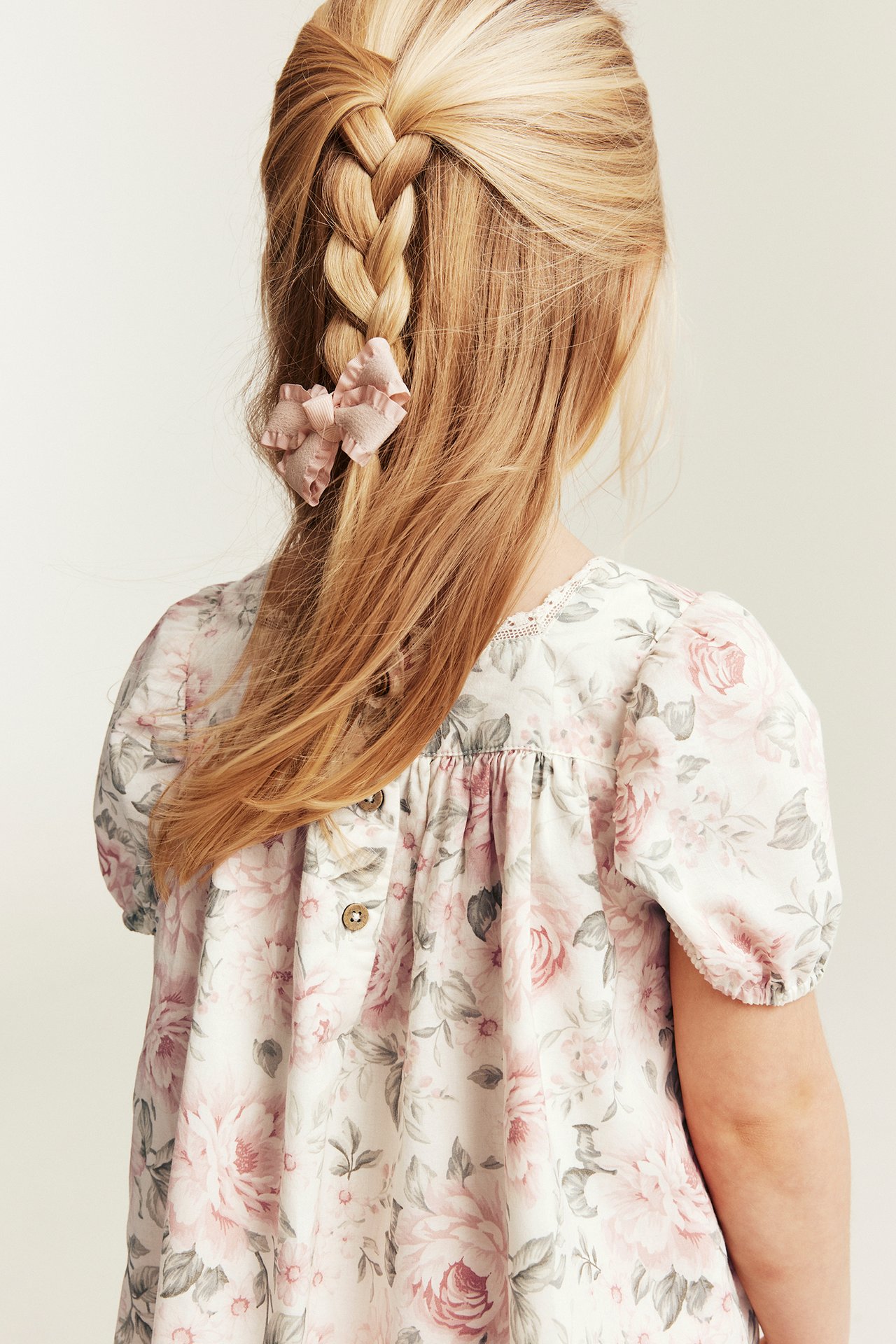 Rückenansicht: Kind trägt cremeweißes florales Baumwollkleid mit Rückenknöpfen, Puffärmeln. Rosa Rüschenschleife im Haar.