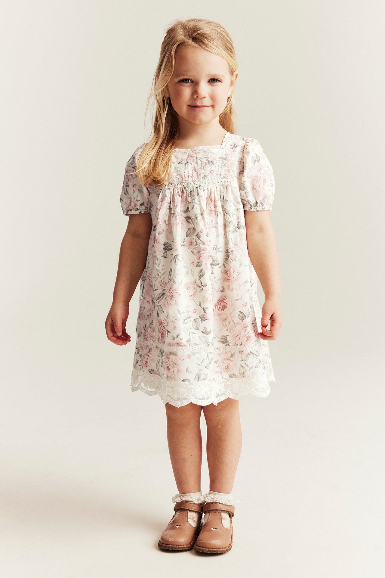 Vorderansicht: Kind in off-weißem Blumen-Baumwollkleid mit Puffärmeln, Spitze und braunen Spangenschuhen.