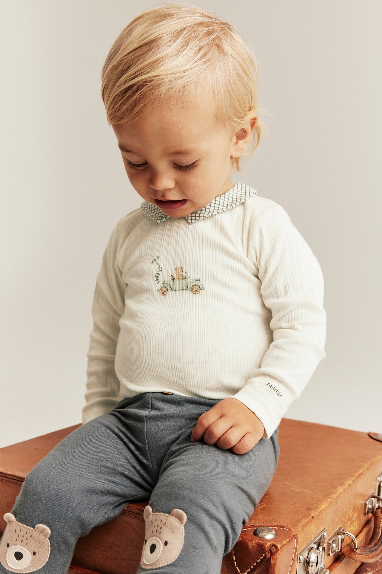 Baby in white ribbed long-sleeve bodysuit with patterned collar, car print, and grey bear-patch leggings.