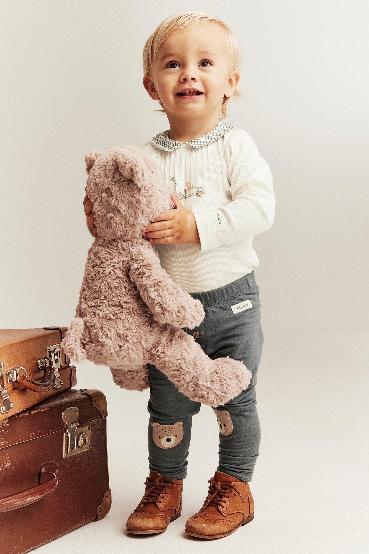 Front view: Baby wears a white ribbed collared bodysuit, grey leggings with bear patches, brown boots, holding a plush bear.