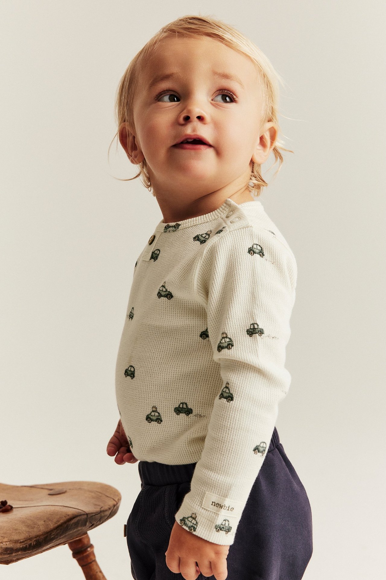 Seitenansicht eines Babys in offwhite Waffelstrick-Body mit grünem Automuster und dunkelblauer Hose.