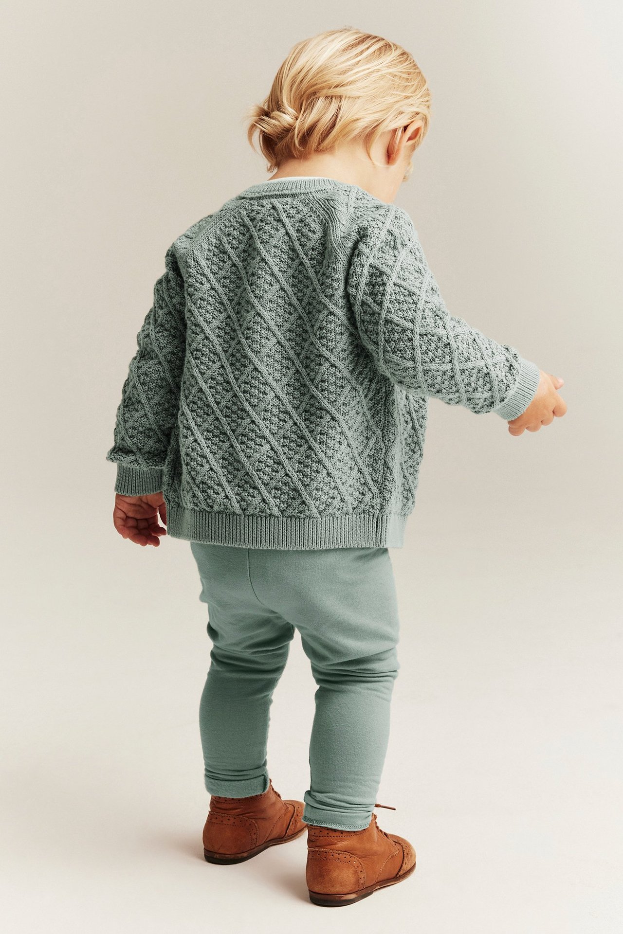 Back view of baby in light green brushed leggings, a matching knitted cardigan, and brown lace-up shoes.