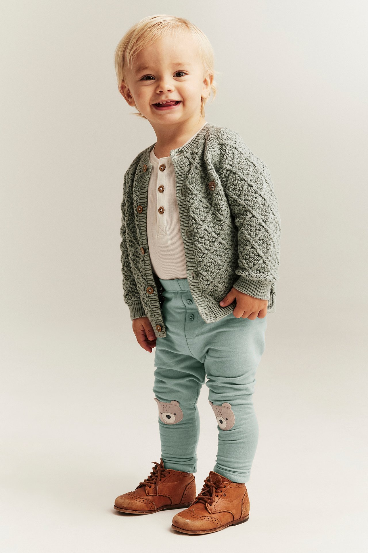 Front view: baby wears light green leggings with bear knee patches, a white Henley, and grey-green cardigan.