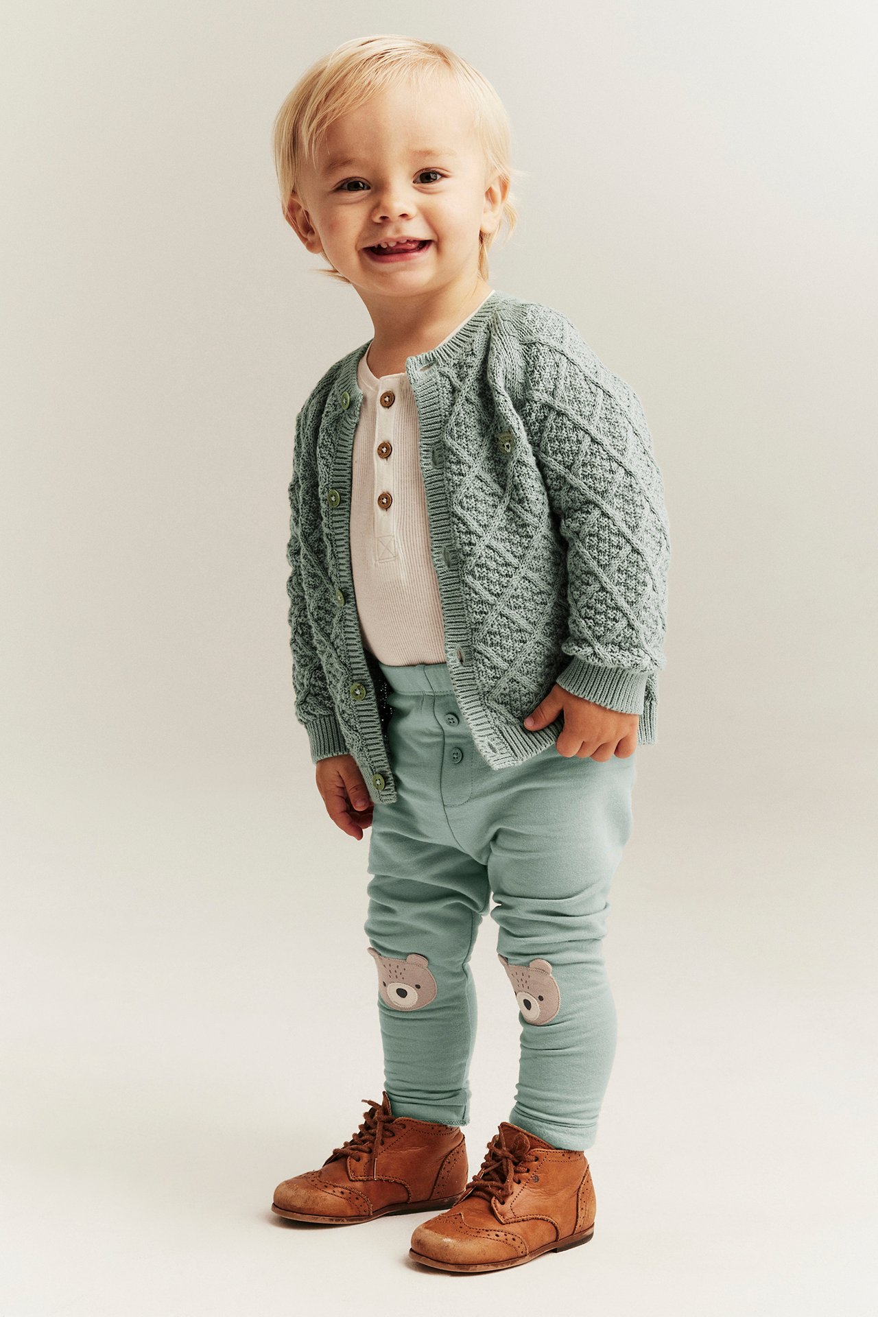 Baby in light green leggings with bear appliques, a cream henley, green cardigan, and brown lace-up boots.