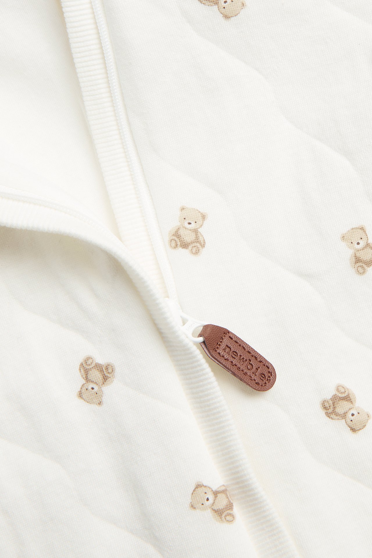 Close-up: off-white quilted baby overall with teddy bears, hidden zipper, and brown pull tag.