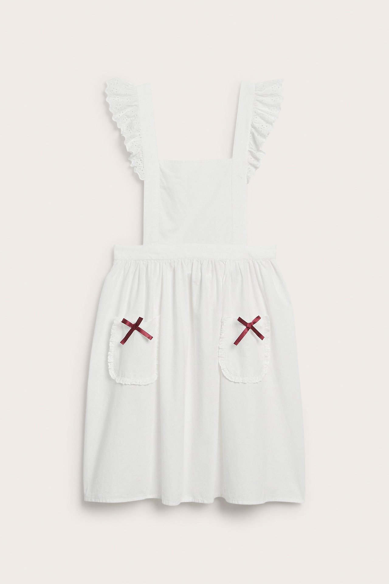 Front view of an off-white cotton apron with ruffled lace straps and red bows on the pockets.