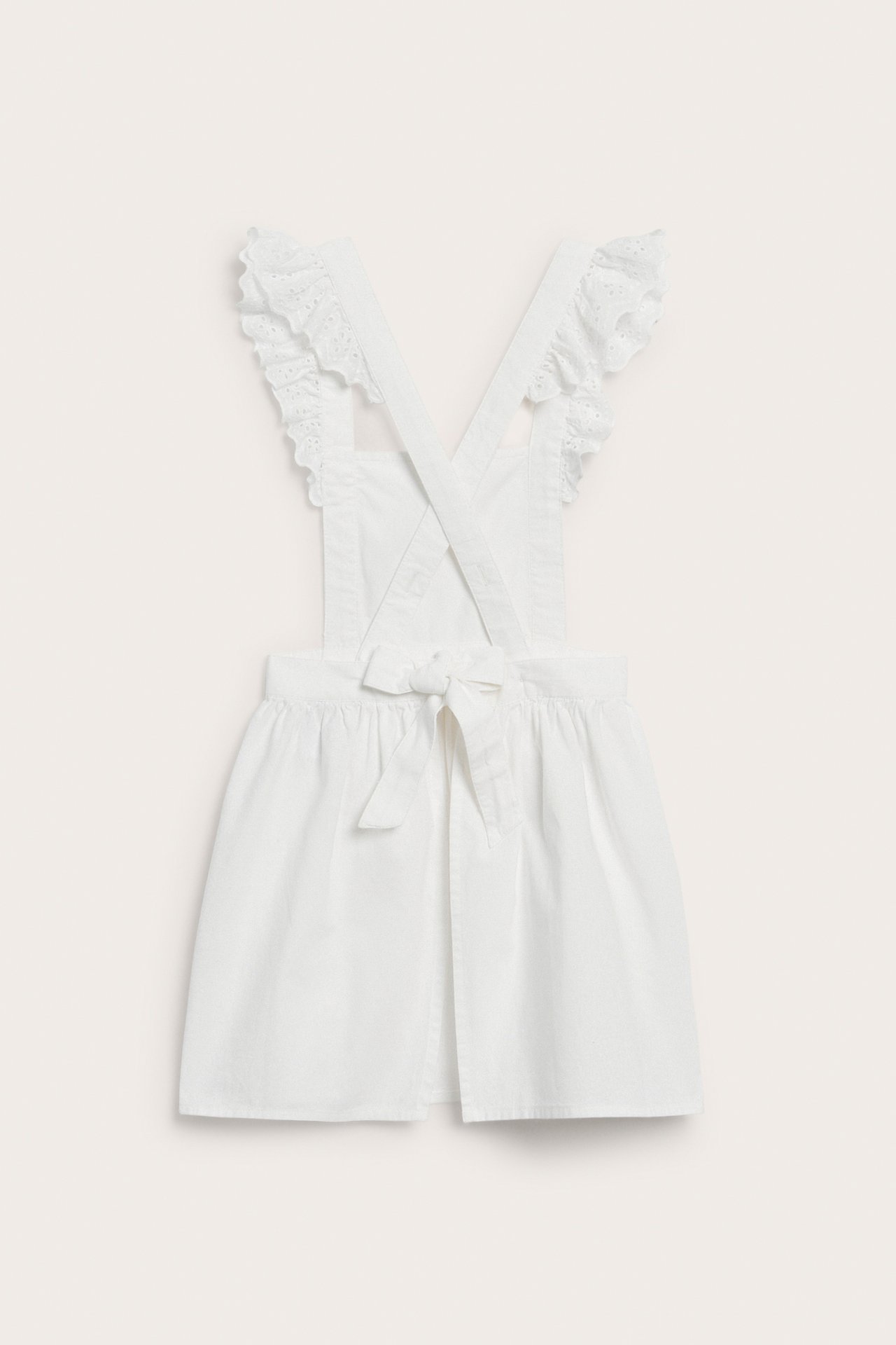 Back view of a kid's off-white cotton apron dress with ruffled straps and a tied back bow.