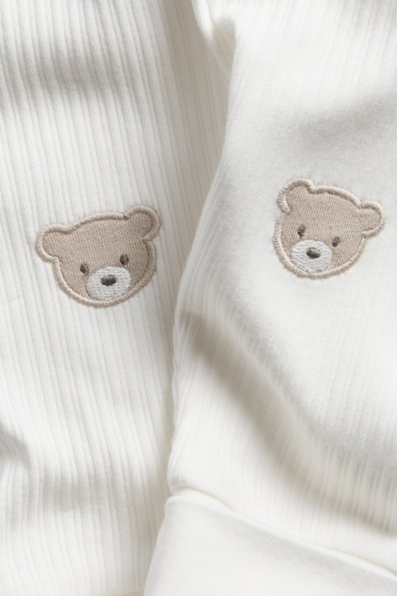 Nahaufnahme von zwei weißen, gerippten Babykleidungsstücken mit hellbrauner Bärenstickerei.