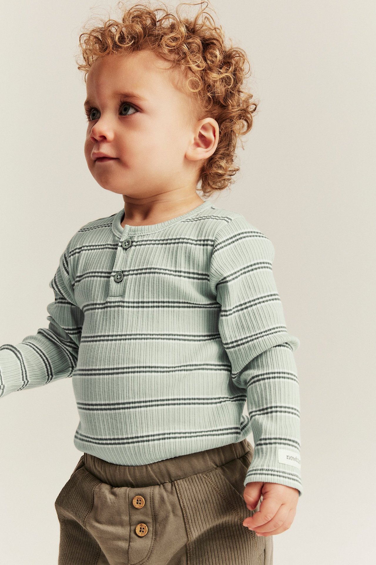 Front view of a baby wearing a light green ribbed long-sleeved striped bodysuit with buttons and brown trousers.