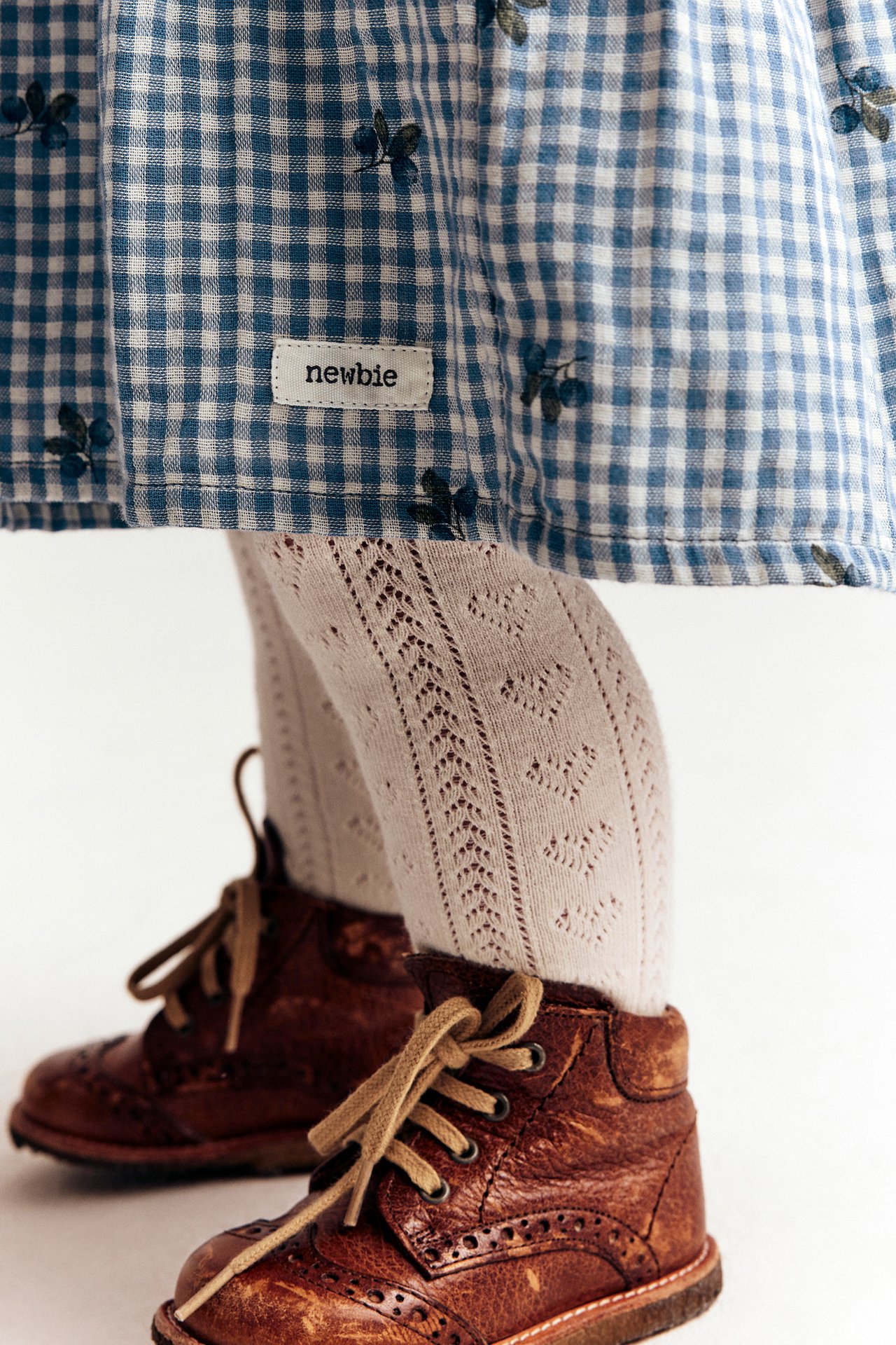 Close-up of a kid wearing a blue checkered cotton dress with blueberry print, light patterned tights, and brown lace-up boots.