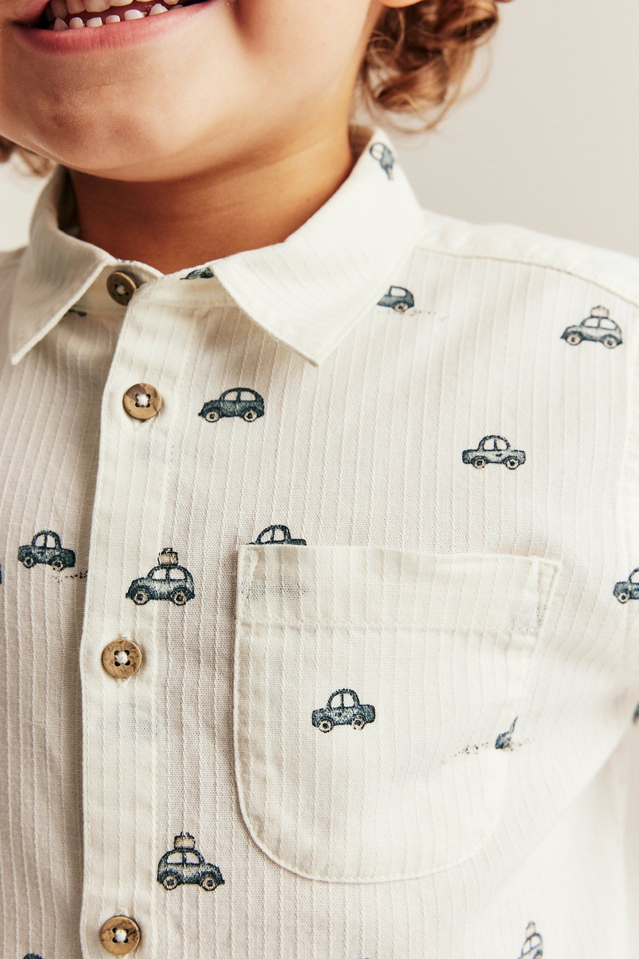 Close-up, front view: kid's off-white ribbed cotton shirt with dark car pattern, wooden buttons, chest pocket.