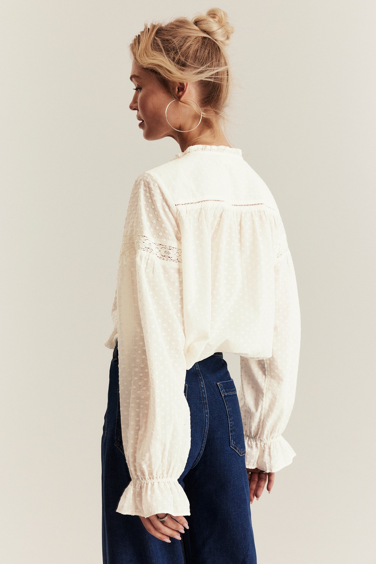 Side view: Woman in off-white woven blouse with lace, ruffled details, dark blue jeans, and hoop earrings.