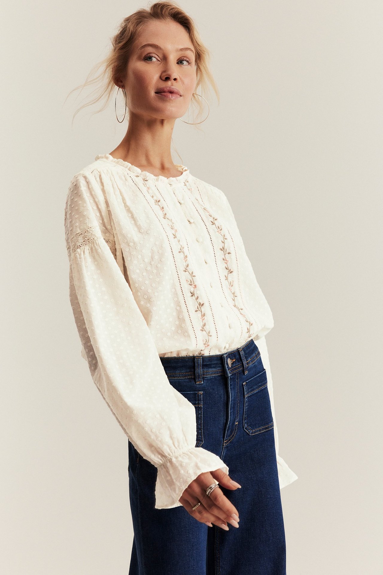 Front view of a woman wearing an off-white embroidered blouse with ruffles, blue jeans, silver hoop earrings, and rings.
