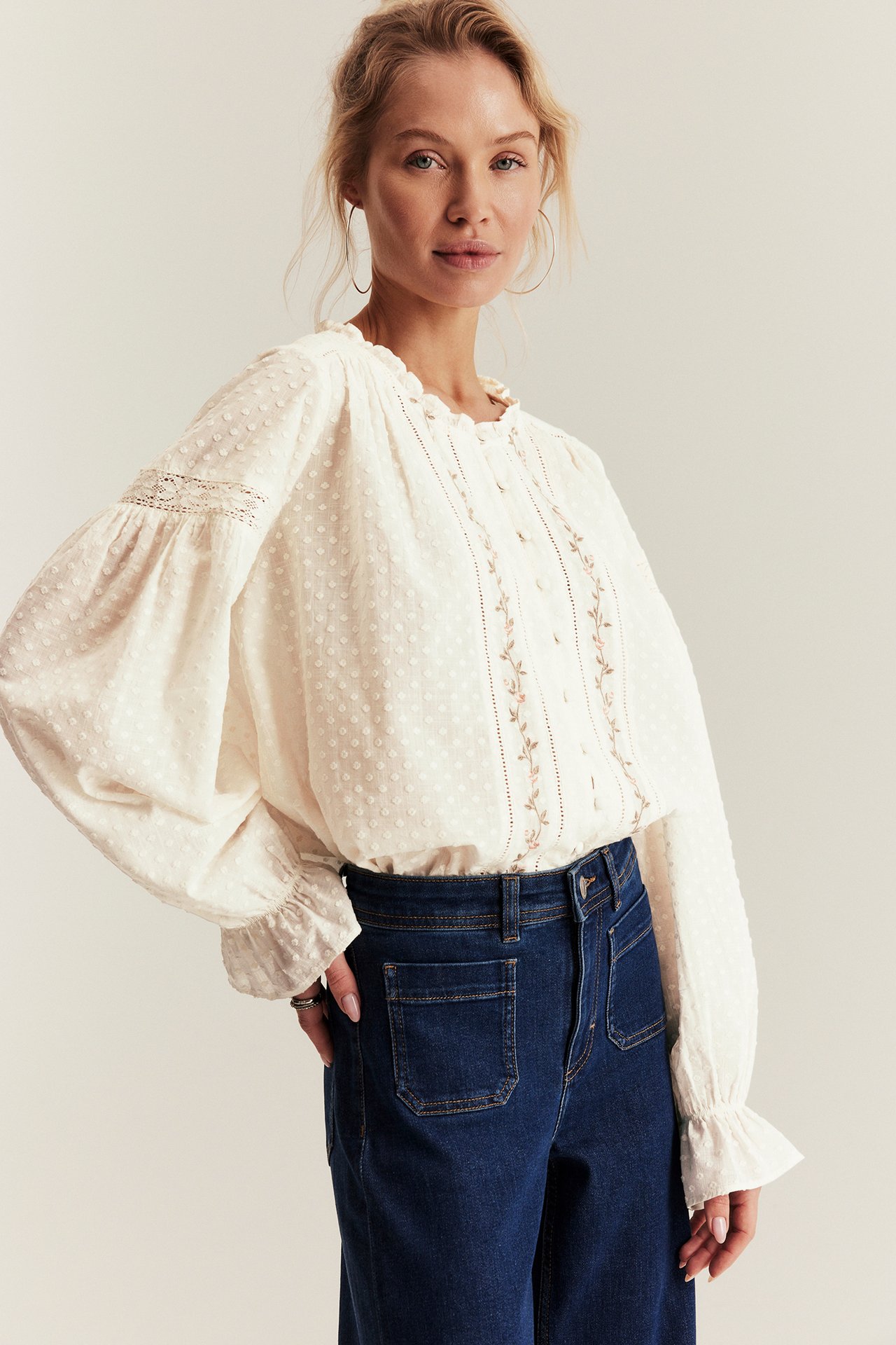 Front view of a woman in an offwhite woven blouse with embroidery, lace inserts, and dark blue jeans.