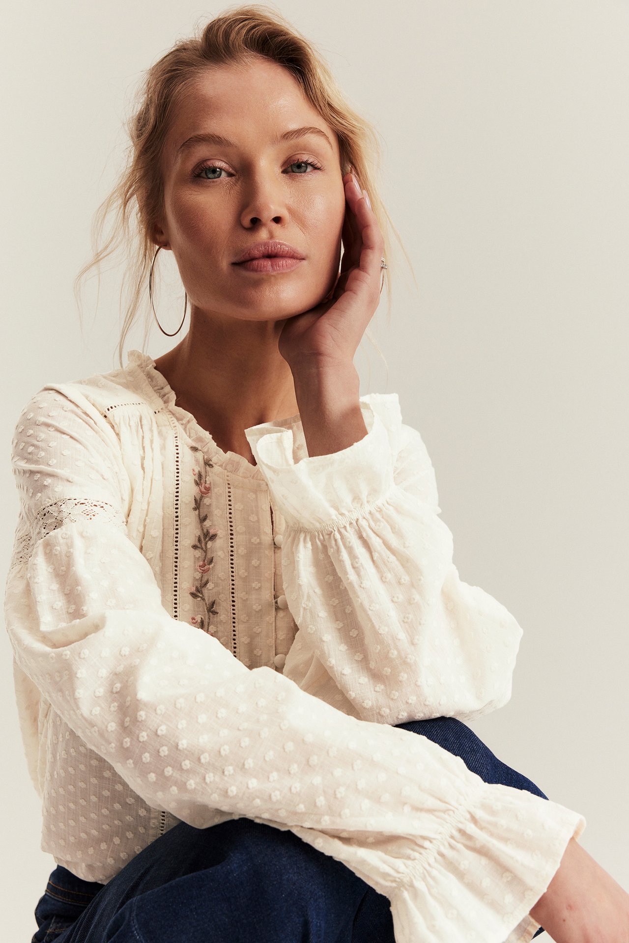 Front view: Woman in off-white cotton blouse with floral embroidery, lace, ruffled collar, cuffs. Wears dark jeans.
