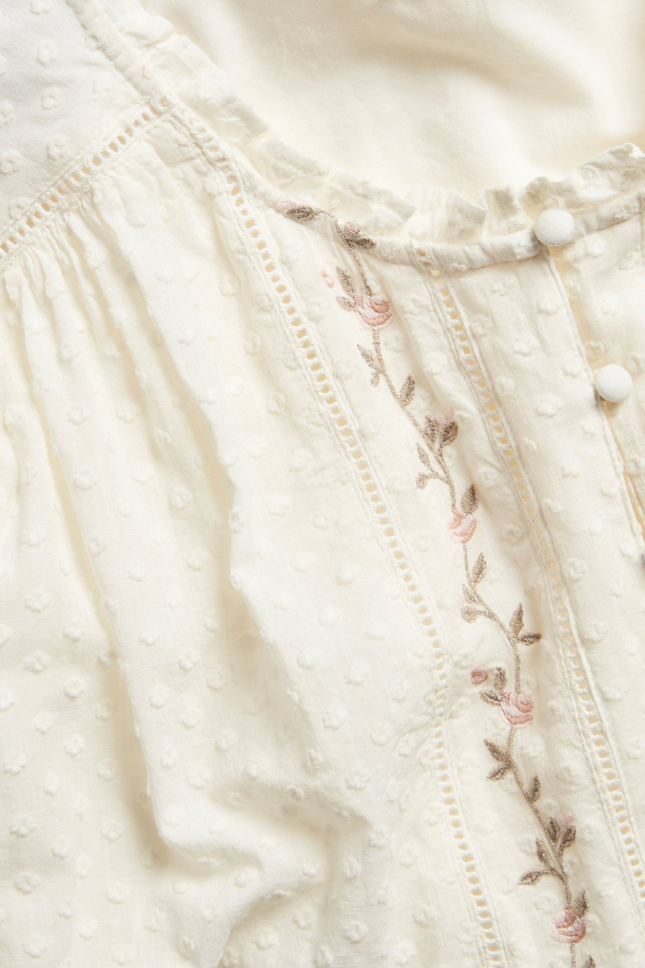 Close-up: Off-white woman's woven blouse with lace, floral embroidery, dotted fabric, and covered buttons.