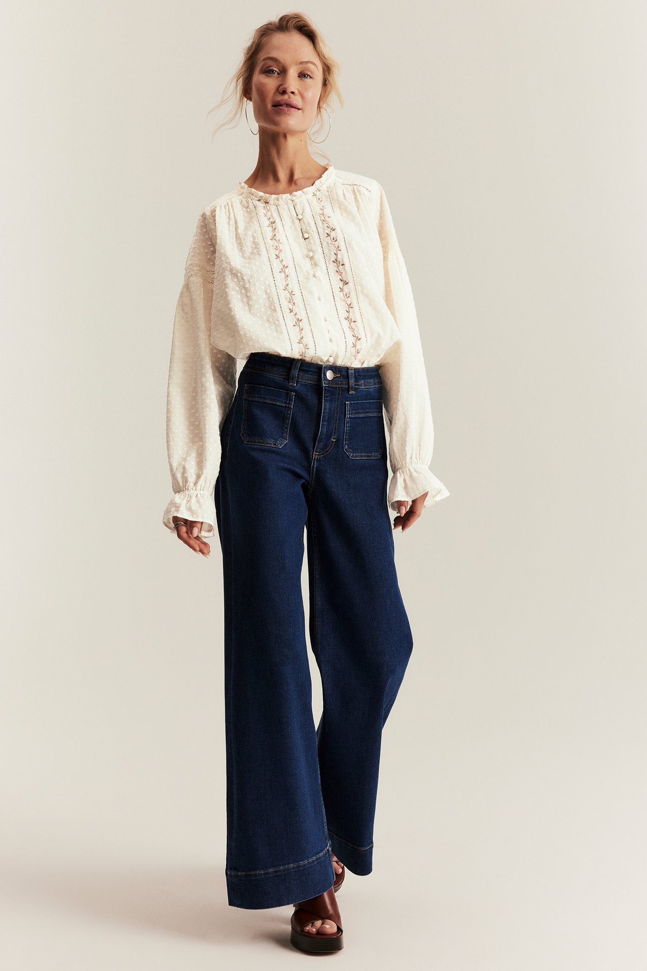 Front view: Woman in off-white embroidered cotton blouse, dark blue wide-leg jeans, and brown sandals.