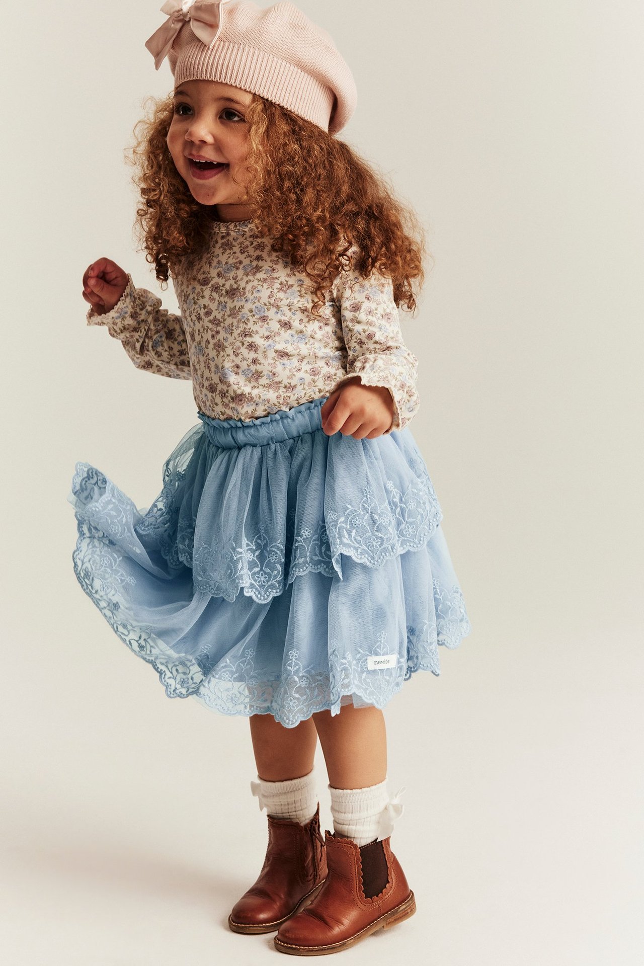Front view of a kid in a blue embroidered mesh ruffle skirt, a floral top, pink beret, and brown boots.