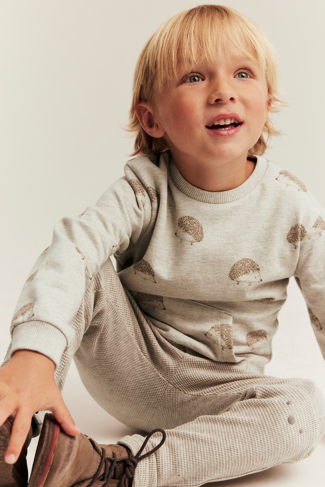 Front view of a kid in a beige hedgehog-patterned sweatshirt, beige pants, and brown boots.