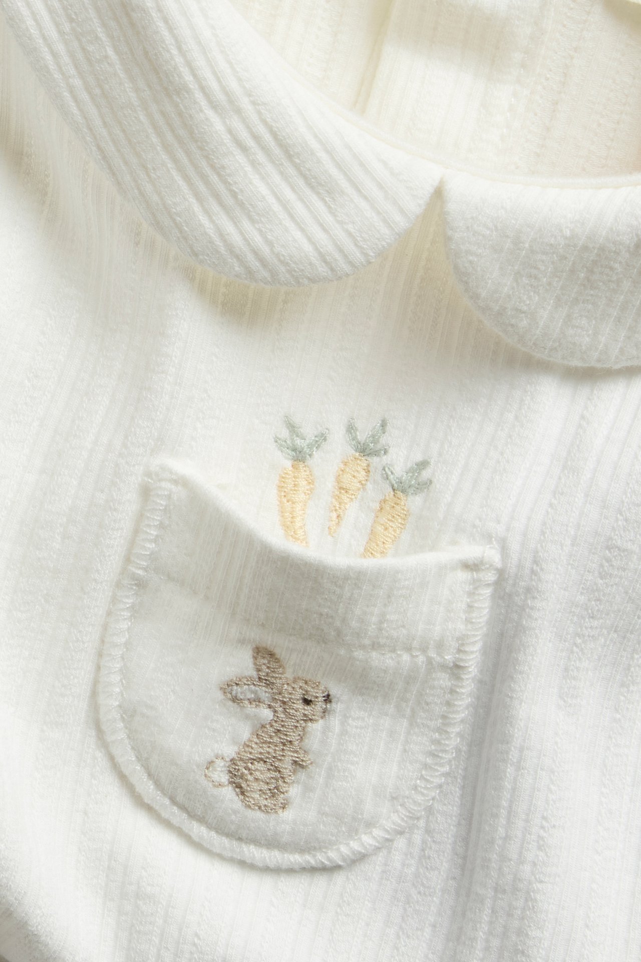 Nahaufnahme eines cremeweißen gerippten Baby-Langarmbodys mit Kragen und bestickter Hasentasche.