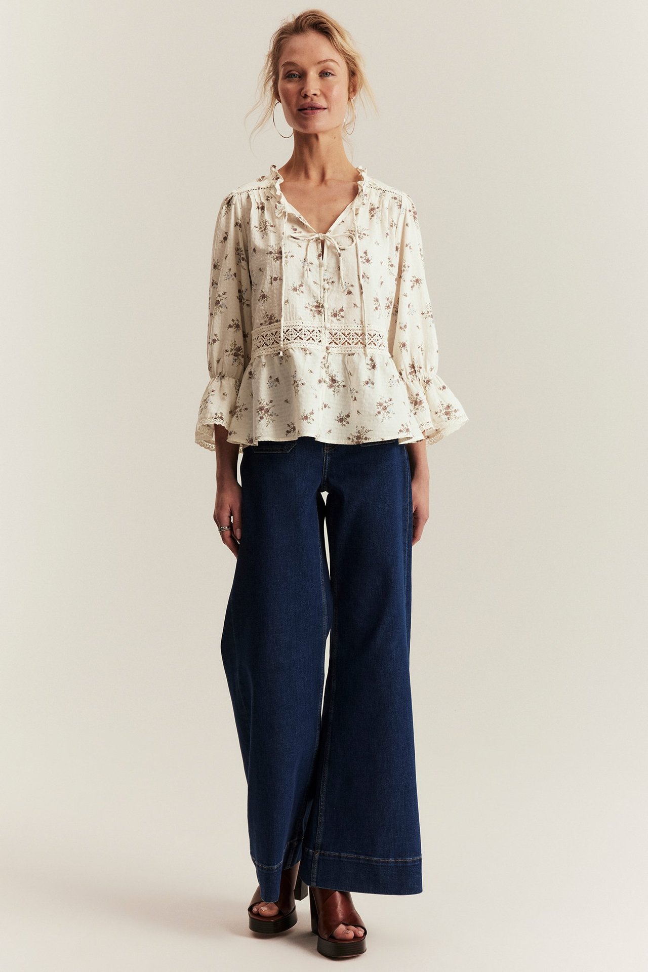 Front view: Woman in off-white floral peplum blouse with lace, dark blue wide-leg jeans and brown sandals.