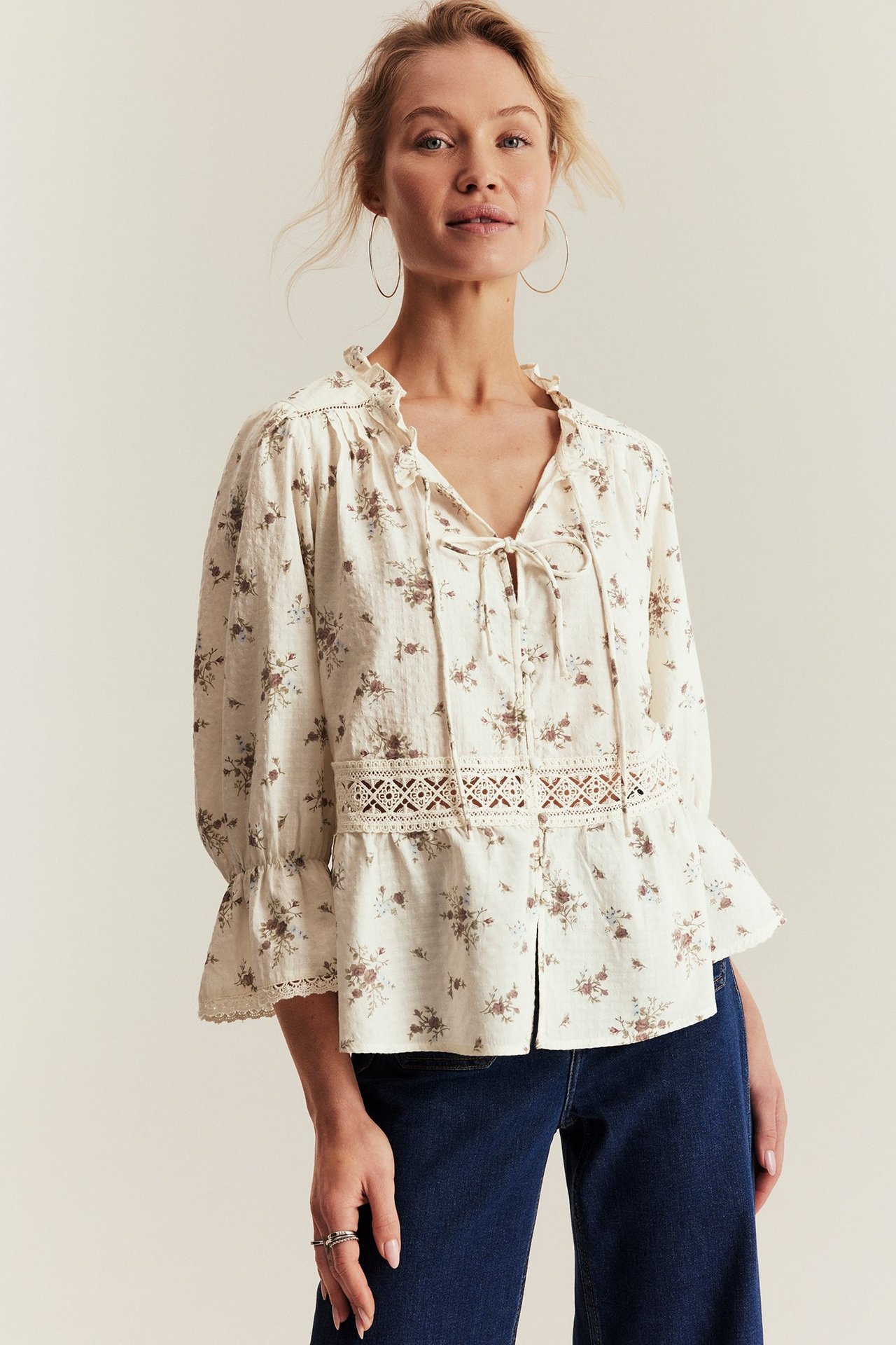 Front view: Woman wears an offwhite floral peplum blouse with lace and 3/4 sleeves, paired with dark jeans.