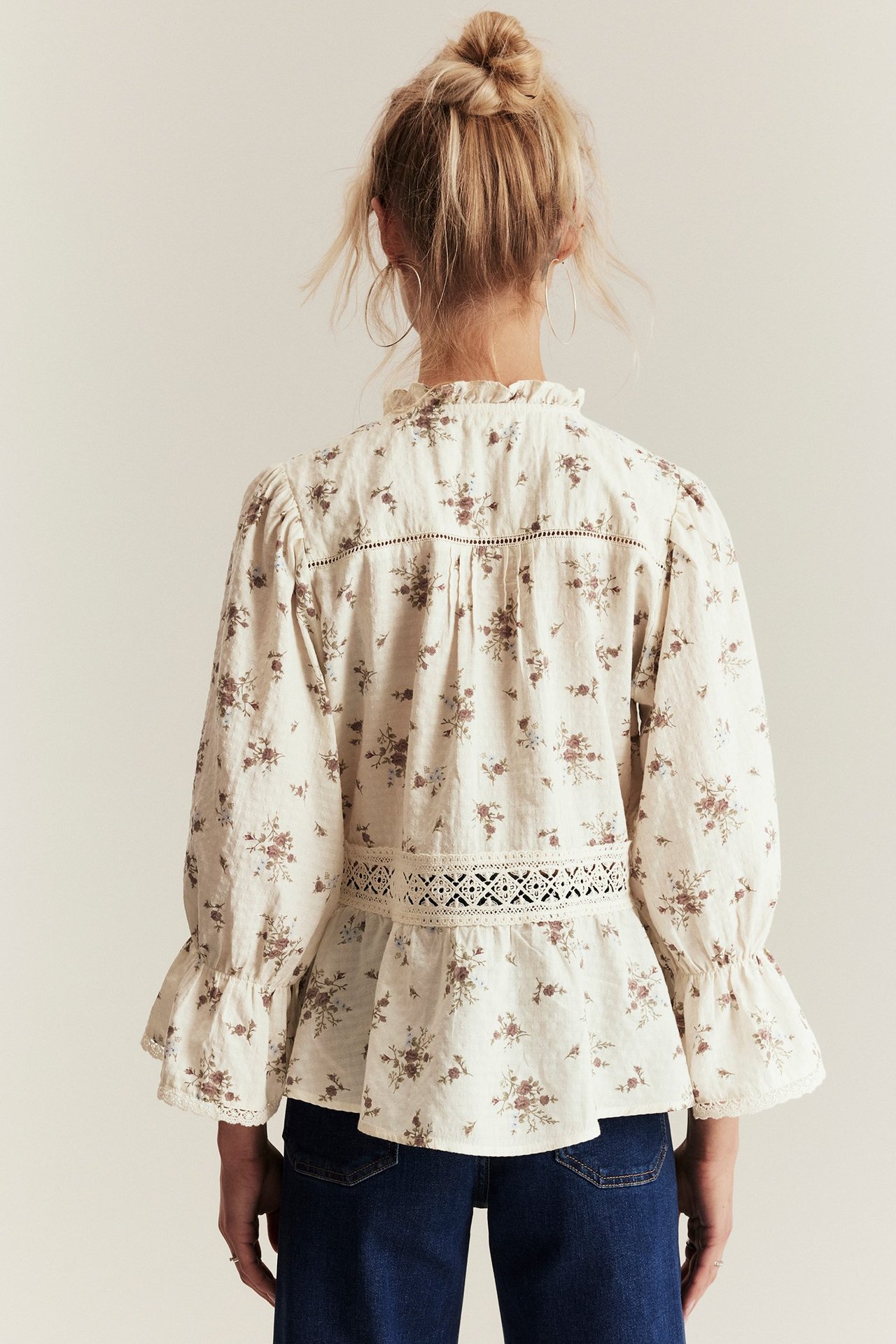 Back view of woman in off-white floral peplum blouse with lace details, ruffled collar, and dark blue jeans.