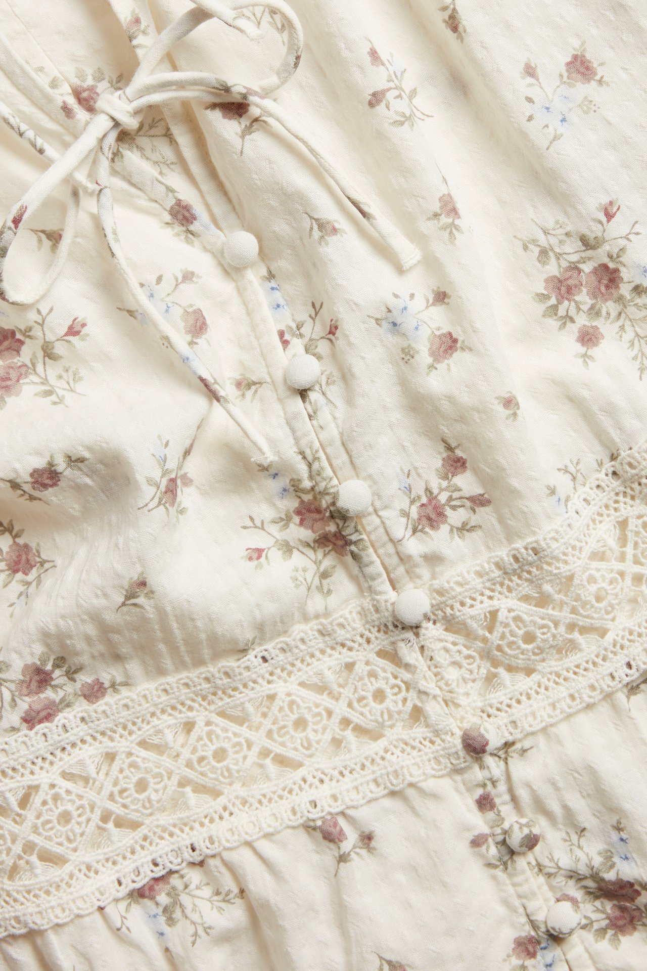 Nahaufnahme einer cremefarbenen Damen-Bluse mit Blumenmuster, Spitze, bezogenen Knöpfen und Bindebändern.