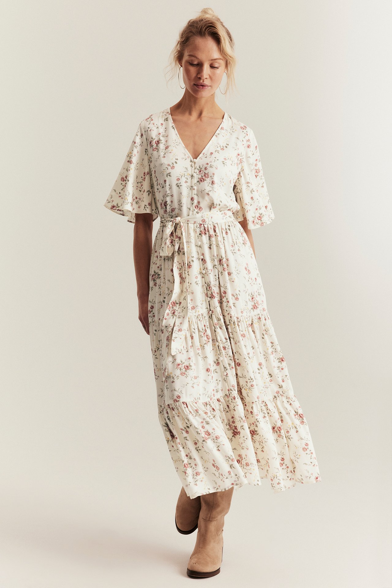 Front view: Woman in off-white floral V-neck maxi dress with short sleeves, tie belt, and brown boots.