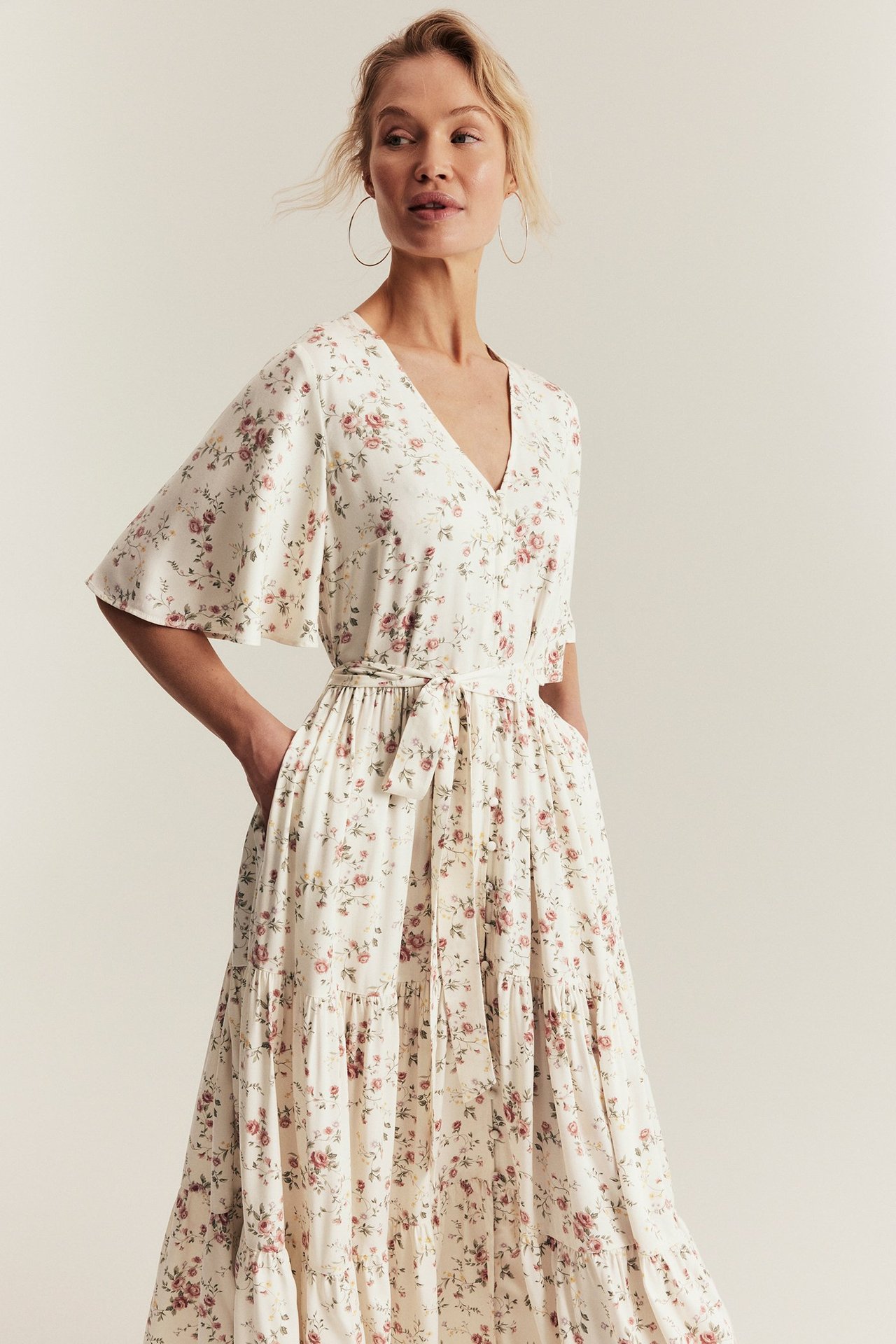 Front-side view: woman in an off-white floral V-neck midi dress with short sleeves and a tie belt.