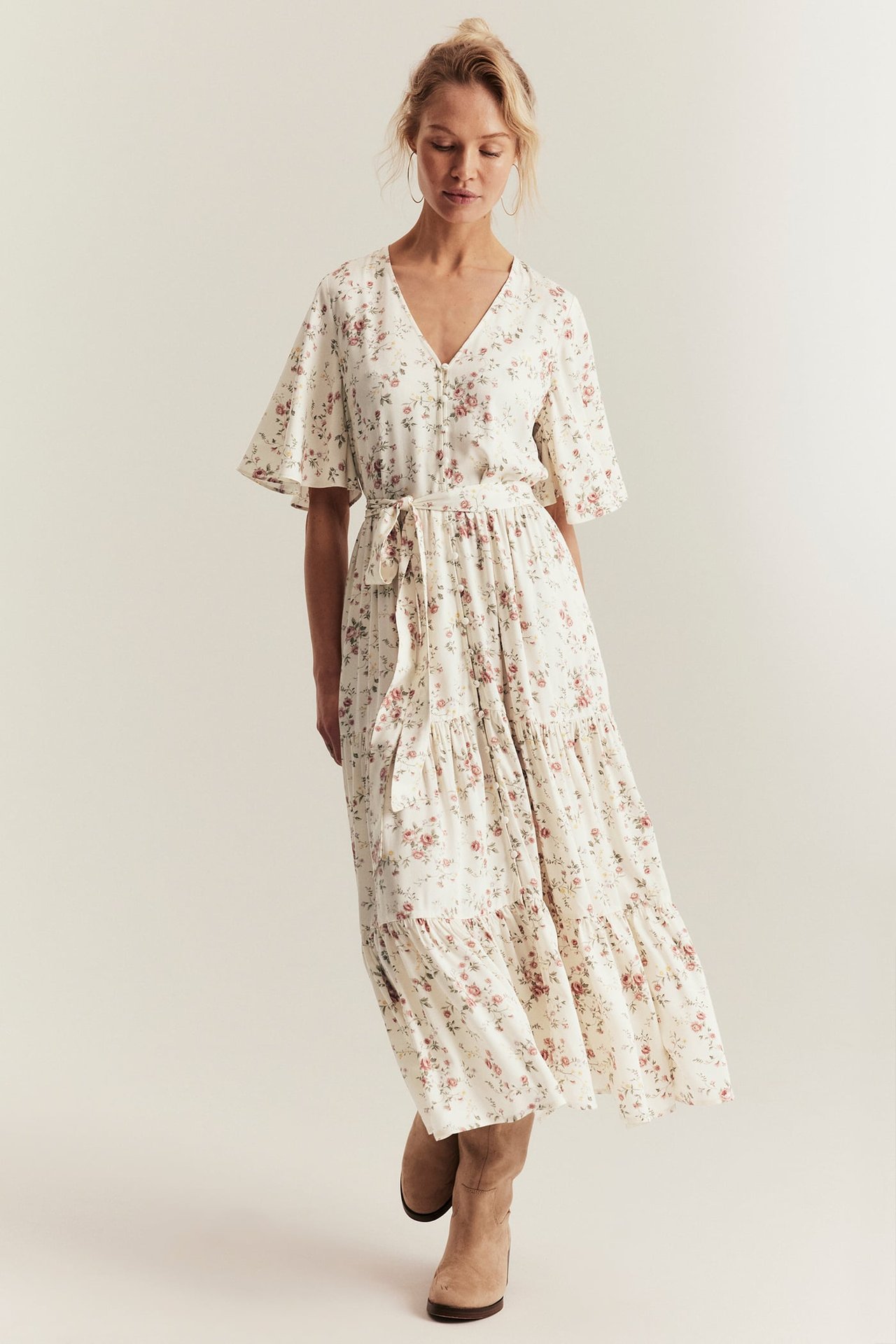 Front view: Woman in off-white floral tiered dress, V-neck, short sleeves, belt, and beige boots.