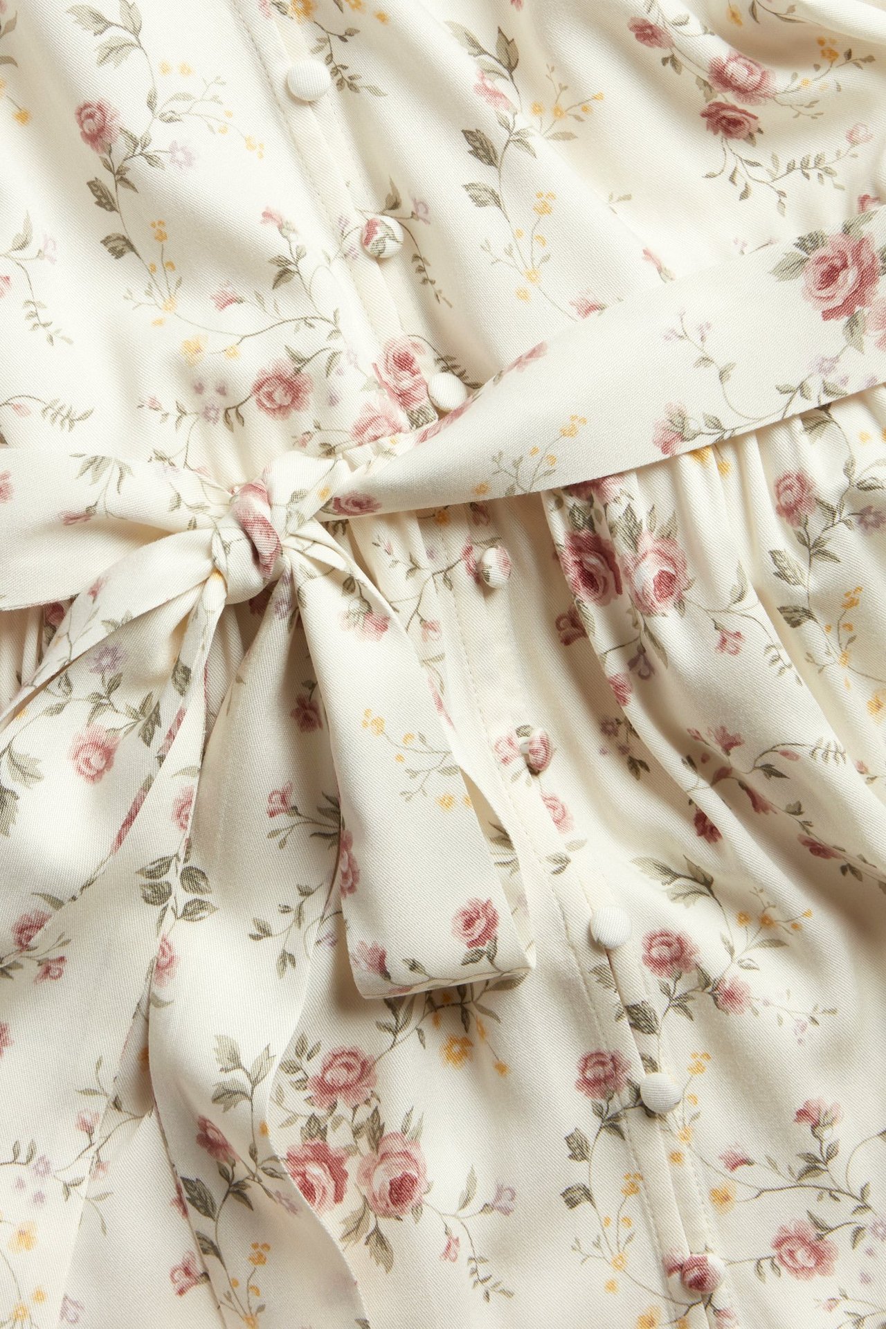 Close-up of an off-white floral viscose dress with pink roses, a tied belt, and covered buttons.