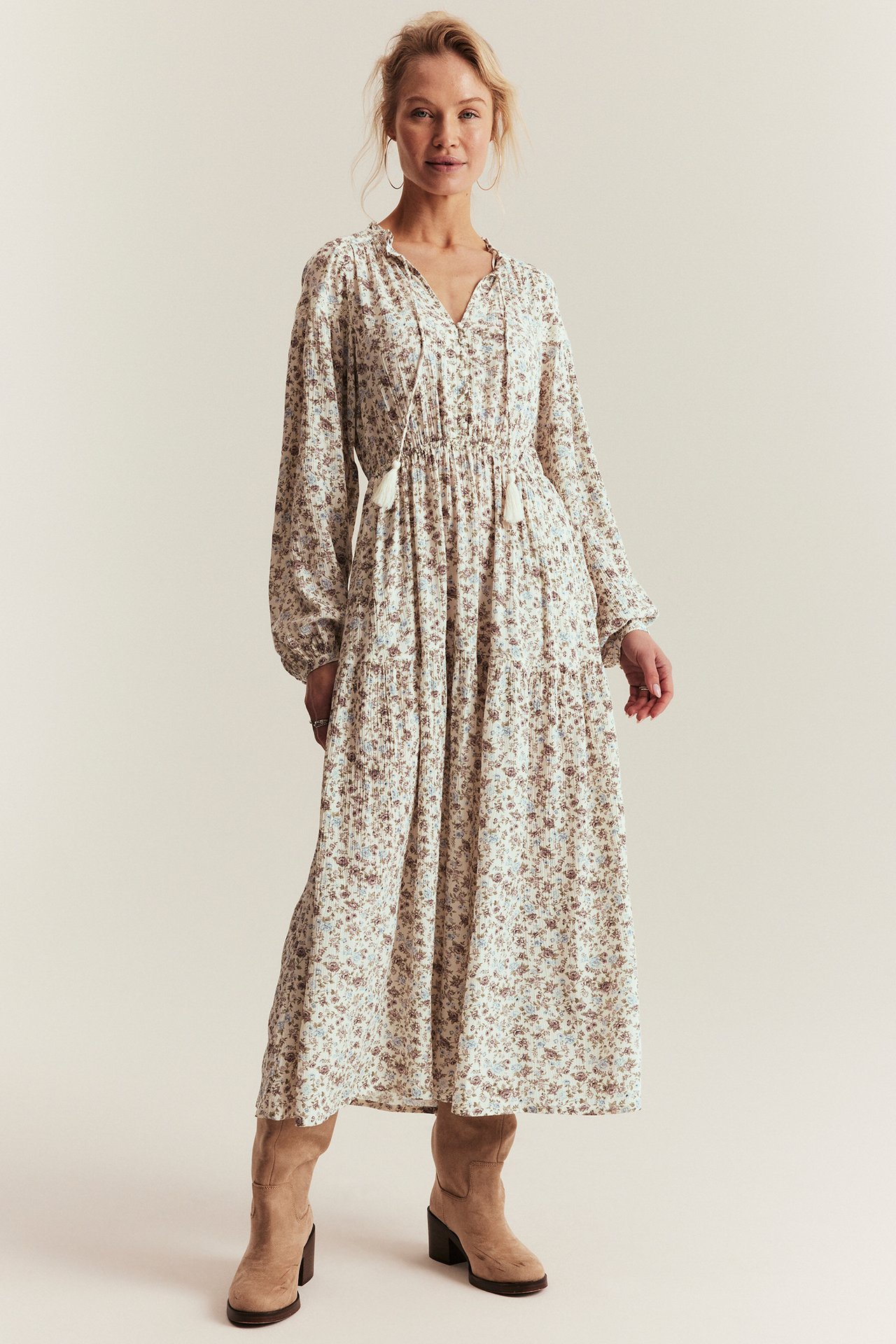 Front view: Woman in off-white floral maxi dress with ruffle collar, tassel ties, long sleeves, and beige boots.