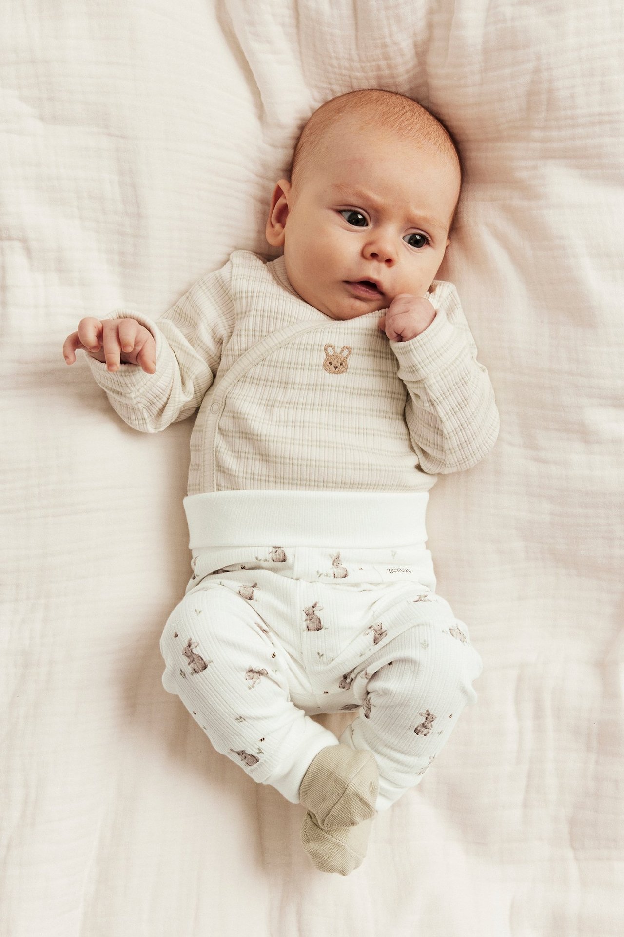 Draufsicht: Baby in off-weißen gerippten Hasen-Leggings, off-weiß gestreiftem Wickelbody und beigen Socken.