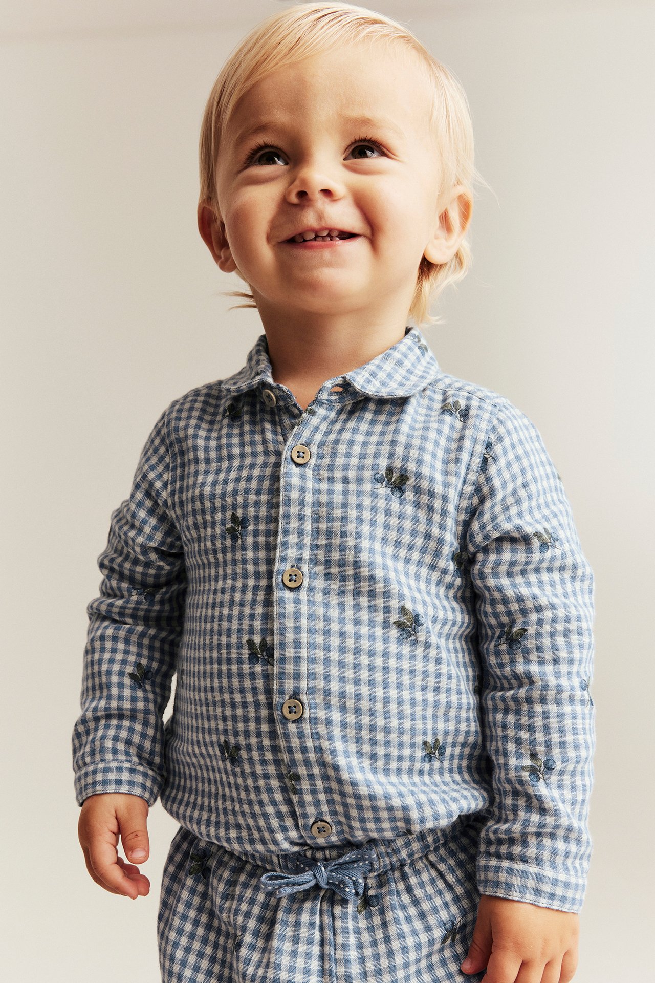 Vorderansicht: lächelndes Baby in einem blau-weiß karierten Baumwoll-Shirtbody mit Heidelbeermuster und Knöpfen.