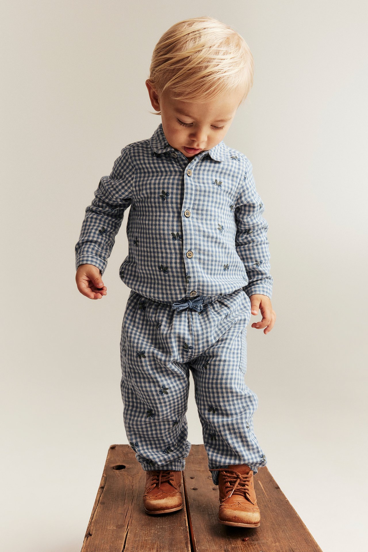 Front view of a kid wearing a blue checked cotton shirt body with blueberry print, matching pants, and brown lace-up shoes.