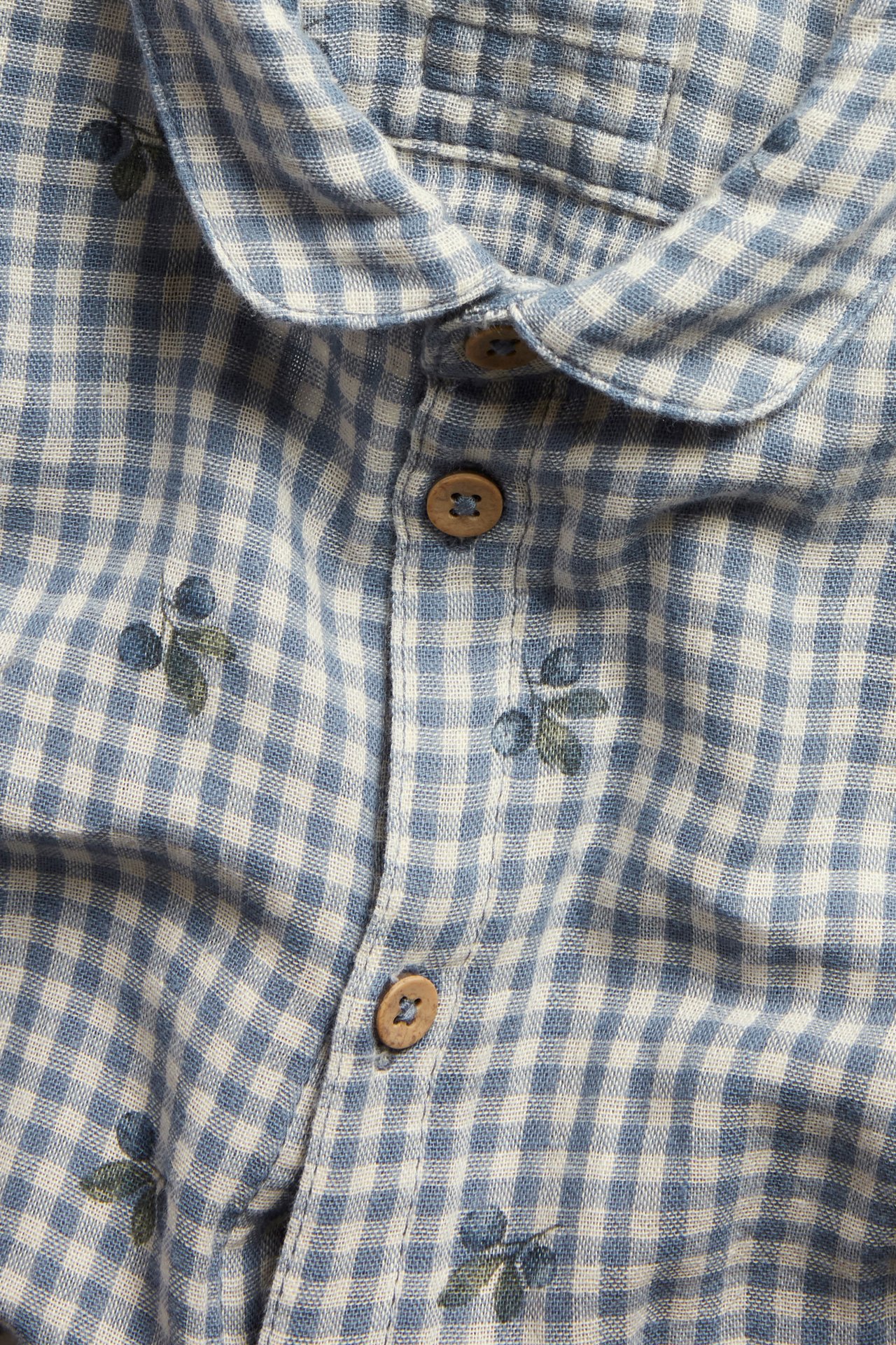 Close-up of a blue and white checked cotton shirt body with collar, wooden buttons, and blueberry print.