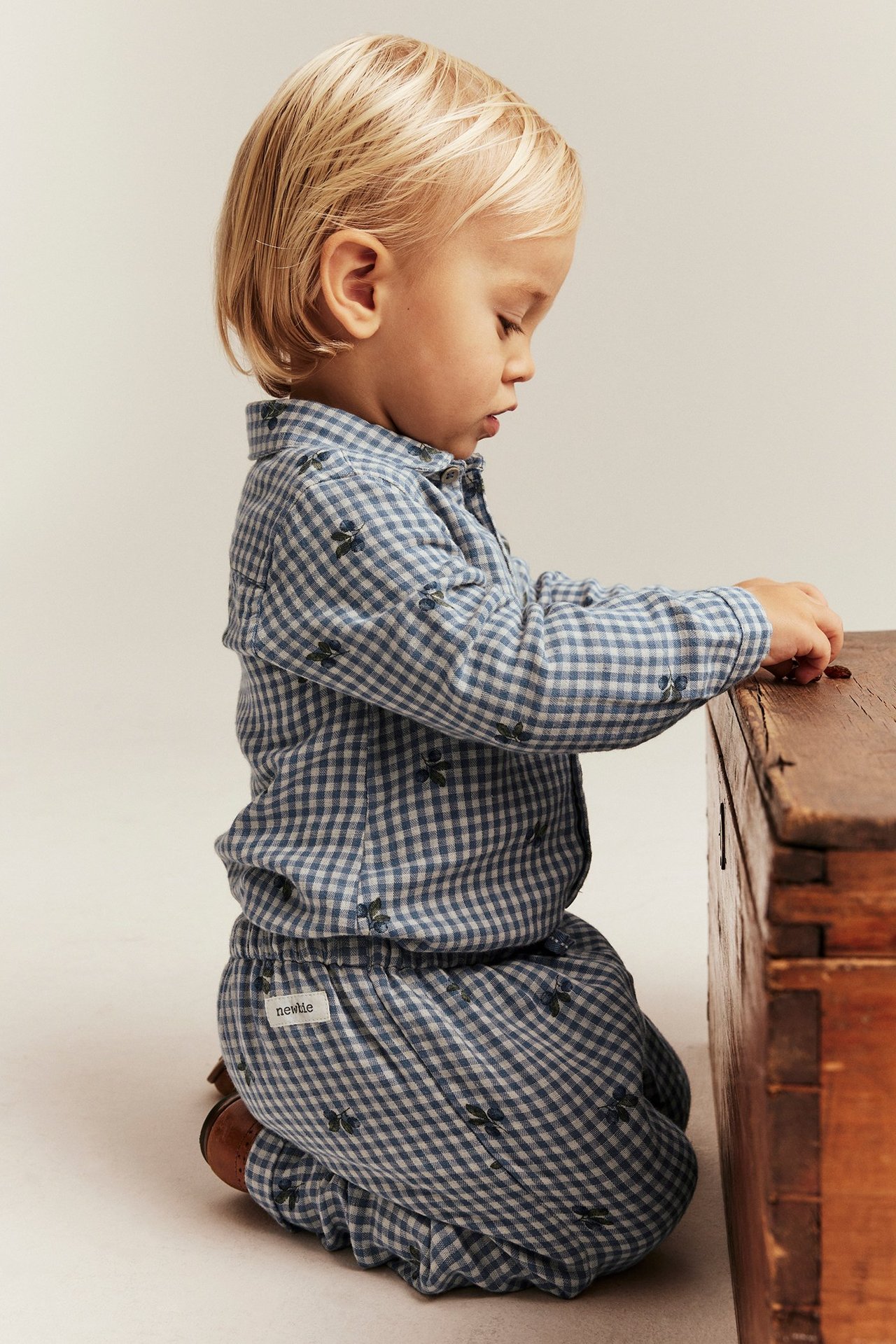 Seitenansicht: Kind trägt blauen, weiß karierten Baumwoll-Shirt-Body mit Heidelbeer-Print, kniet an Holzkiste.