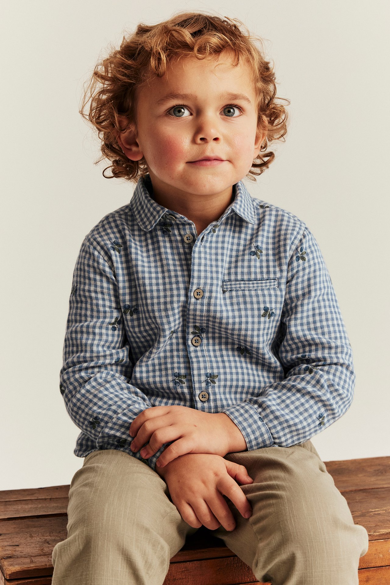 Front view of a kid wearing a blue checkered cotton shirt with blueberry print, chest pocket, and beige trousers.