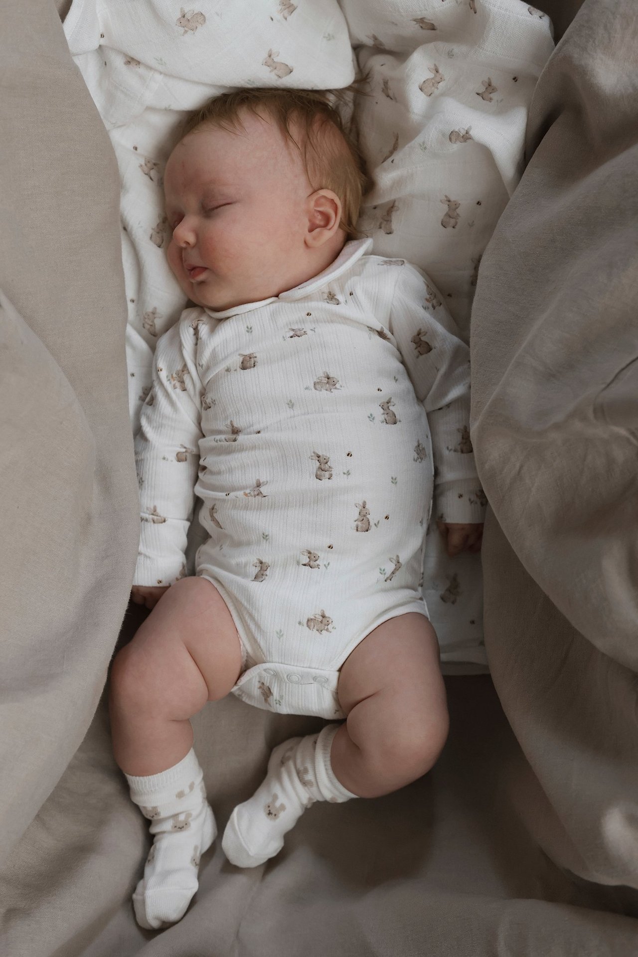 Draufsicht, schlafendes Baby in offweißem Ripp-Langarmbody mit Kaninchendruck und passenden Socken.