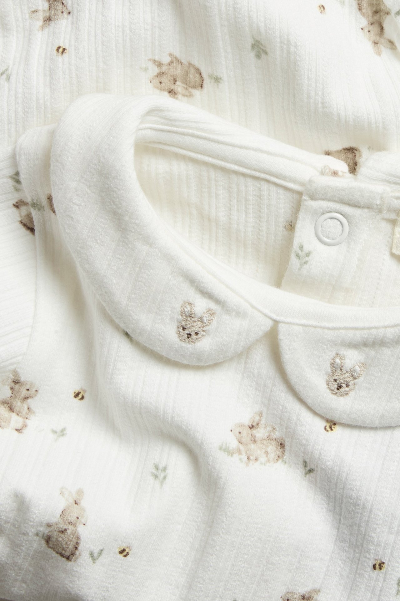 Nahaufnahme eines cremeweißen gerippten Baby-Bodys mit Kragen, bestickten Hasen und Druckknopf am Nacken.