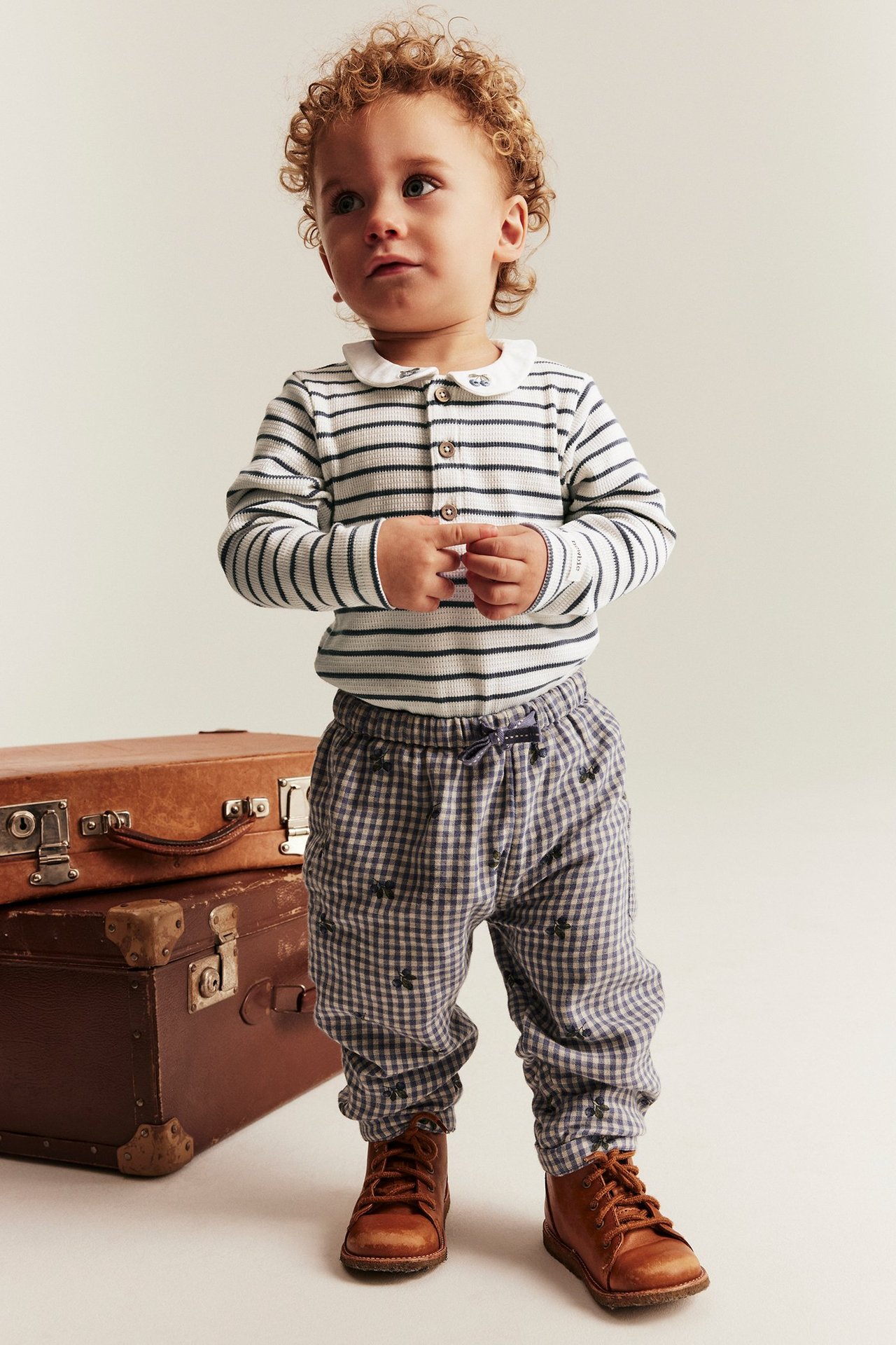 Vorderansicht: Baby in gestreiftem off-white Rippbody mit besticktem Kragen, blaukarierter Hose und braunen Stiefeln.